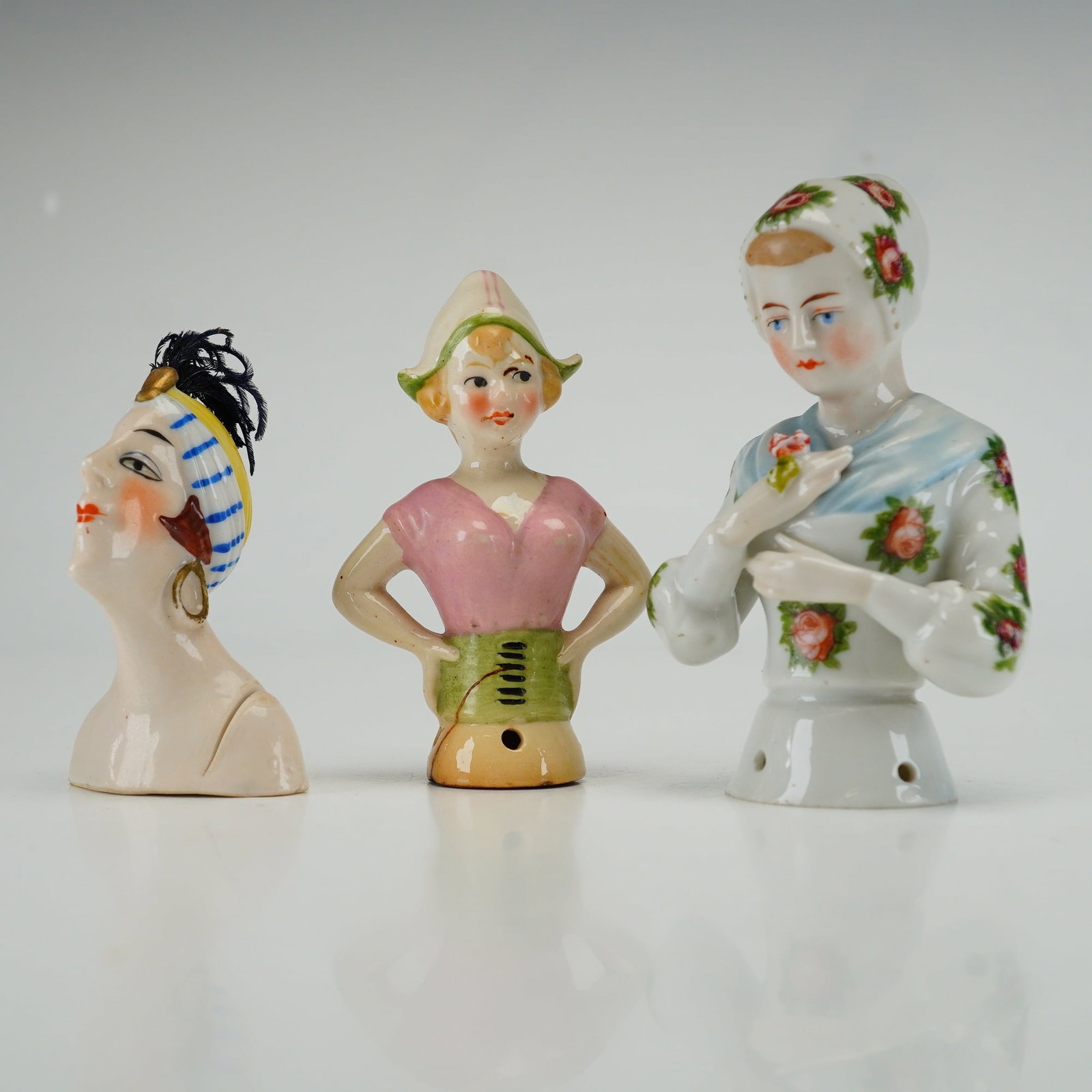 Three Vintage German Style Porcelain Pincushion Half Dolls: These items were manufactured as decorative toppers for pincushions, tea cozies, or whisk brooms. The first figure is an Art Deco style bust of a woman wearing an exotic headpiece with yellow and blue