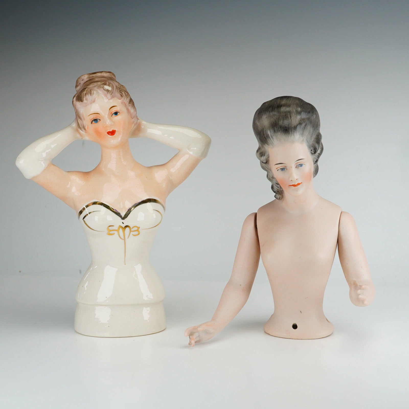 Two Porcelain and Bisque Half Dolls, Japanese and German: This lot consists of two ceramic half dolls. The first is a Japanese glazed porcelain half doll depicting a woman with brown hair in a bun and her arms positioned behind her head. She wears a white st