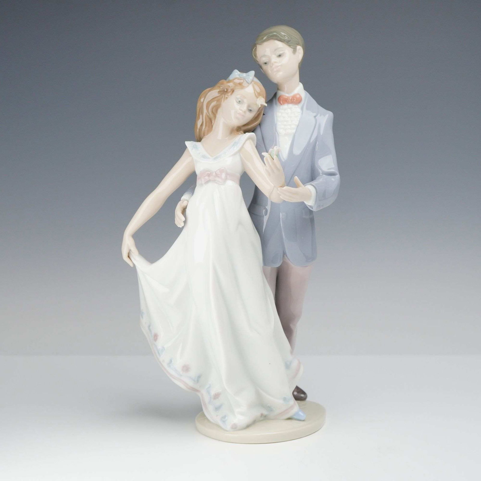 Lladro Glazed Porcelain Figurine Now and Forever 01007642: This Lladro glazed porcelain figurine depicts a young couple in a dancing pose. The woman is dressed in a sleeveless white gown with a pink sash at the waist and a delicate blue and pink floral motif 