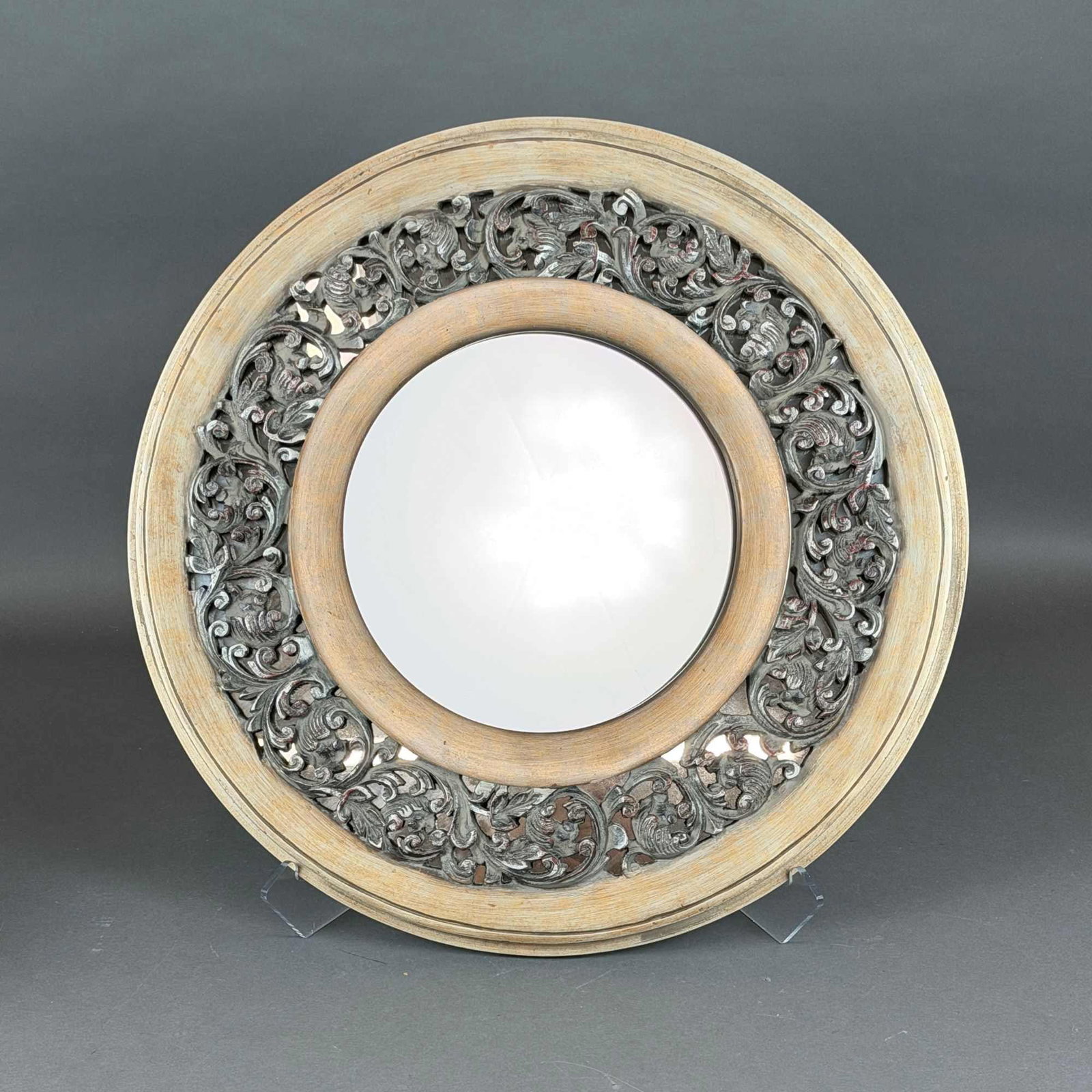 The Bombay Company Bentley Filigree Round Wood Frame Wall Mirror: A contemporary circular wall mirror by The Bombay Company. The wood frame is constructed in three concentric sections. The inner and outer bands feature a distressed silver and pale gold wash over a l