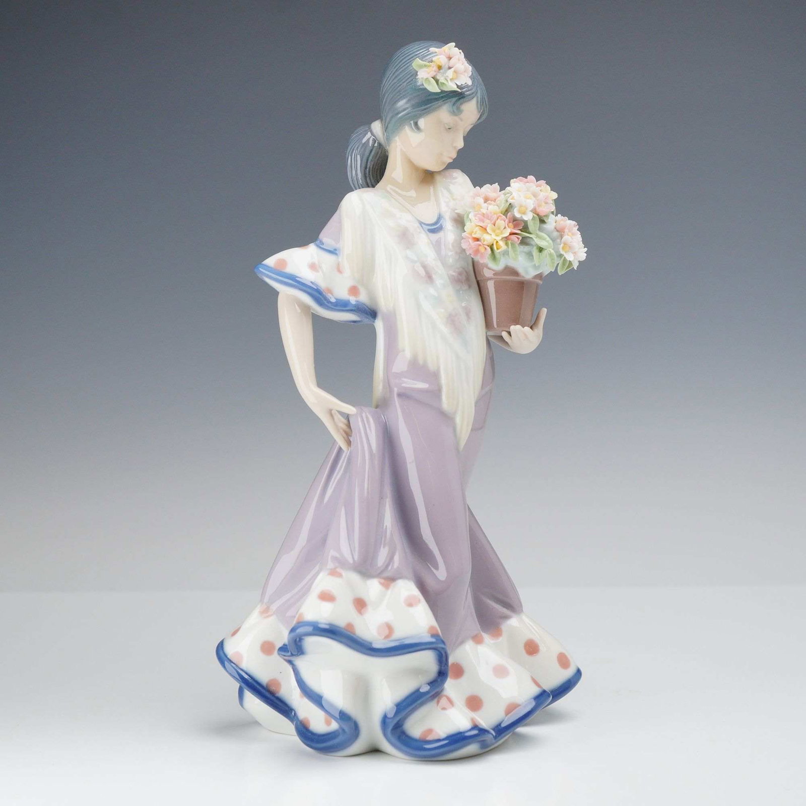 Lladro Glazed Porcelain Figurine, Flor Maria, Number 01005490: This polychrome figurine depicts a young woman in traditional Spanish attire, featuring a lavender-colored gown with a wide, ruffled hem decorated with reddish-orange polka dots and a blue border. A w
