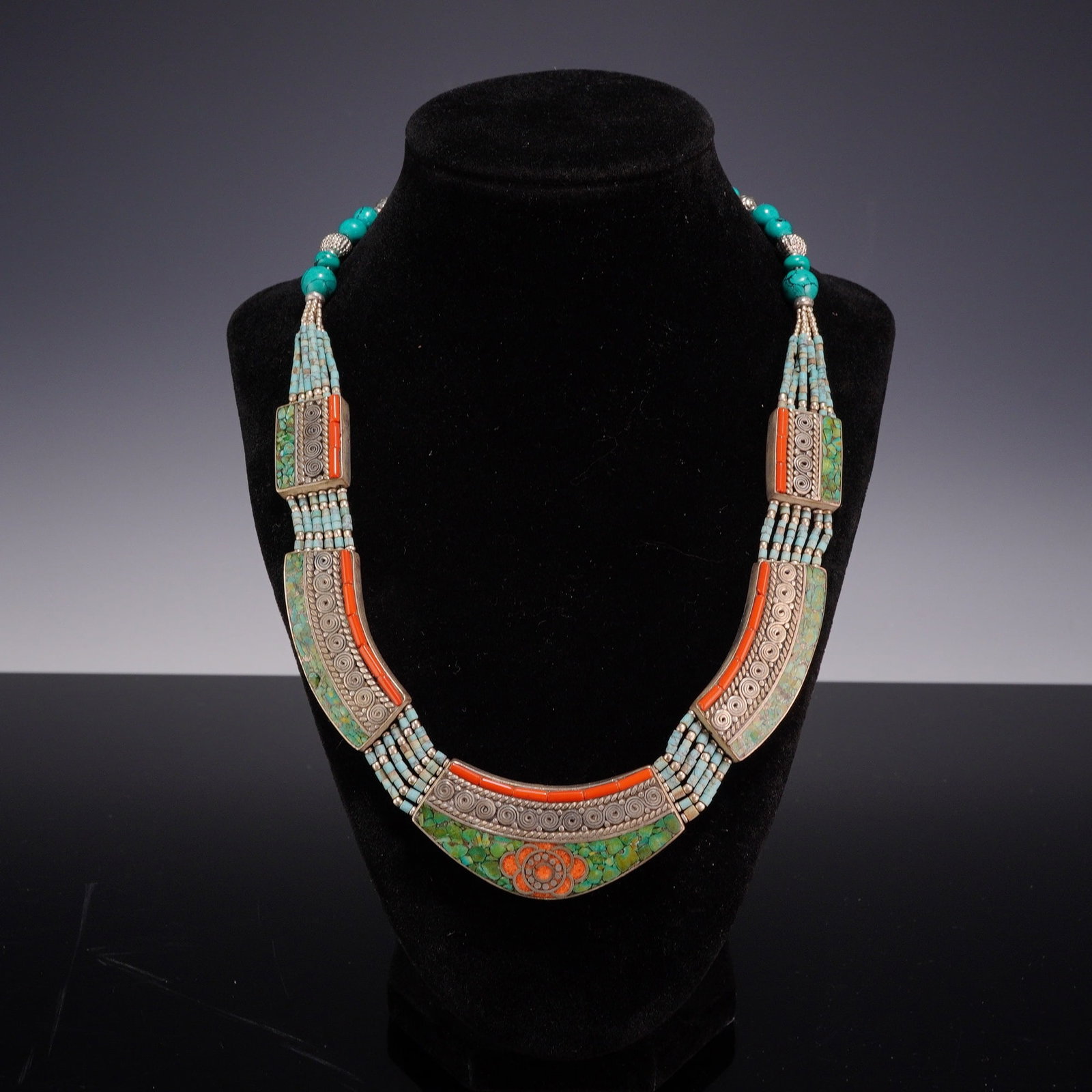 Sterling Silver Tibetan Style Costume Necklace: The bib-style necklace features multiple strands of small turquoise-colored beads and larger round turquoise-colored beads leading to three articulated silver-tone plaques. These plaques are