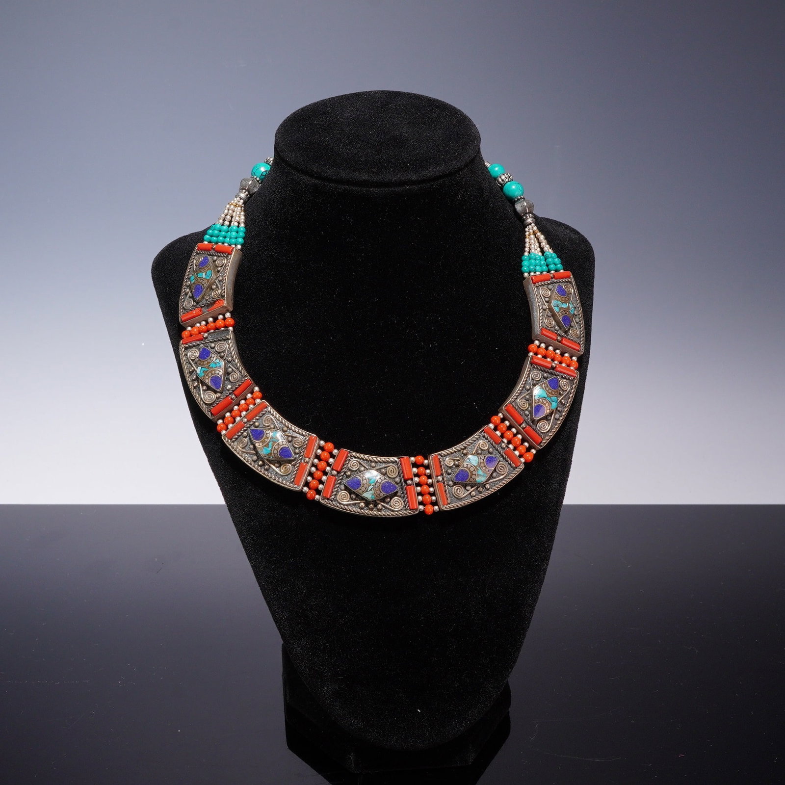 Tibetan-Style Bib Necklace with Turquoise, Lapis Lazuli, and Coral-Colored Accents: This bib-style collar necklace is designed in a traditional Tibetan or Himalayan style. The piece is constructed from silver-tone white metal featuring intricate filigree, wirework spirals, and