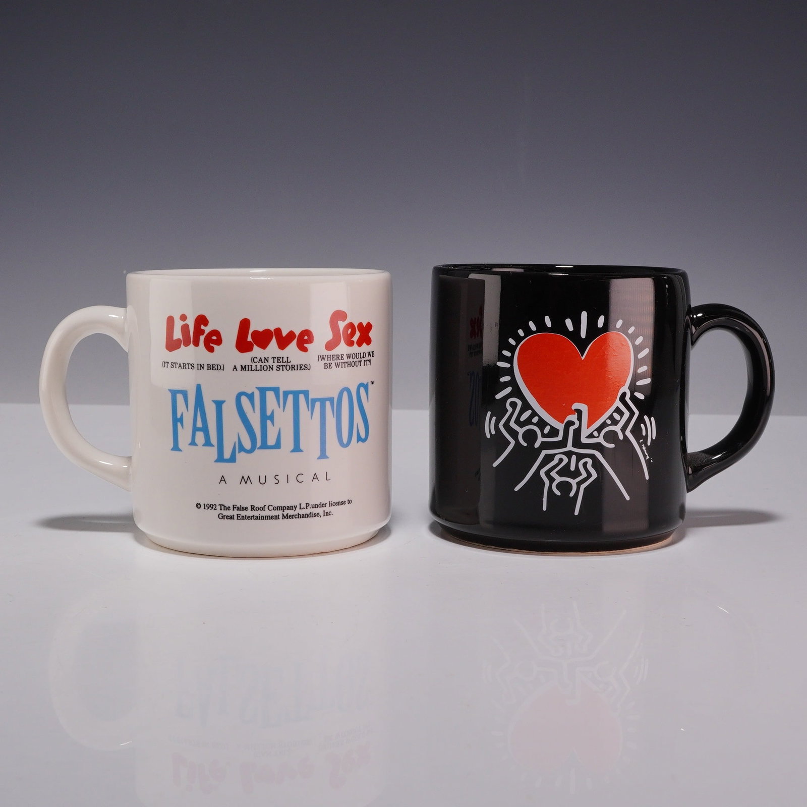 2pc 1992 Falsettos Broadway Musical Mugs with Keith Haring Graphics: A pair of ceramic promotional mugs from the 1992 Broadway production of the musical Falsettos, featuring graphics by artist Keith Haring. The collection includes one white glazed mug and one black