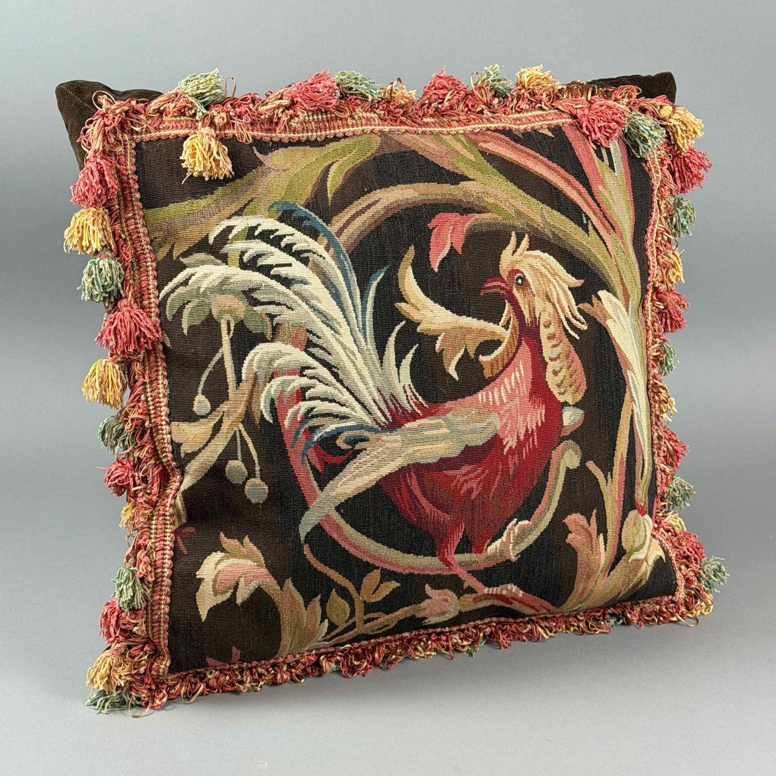 Handwoven Aubusson Style Rooster Design Throw Pillow with Tassel Fringe: This handwoven Aubusson throw pillow features a decorative tapestry face depicting a stylized rooster or cockerel. The bird is rendered in shades of red, cream, and tan, set against a dark brown groun