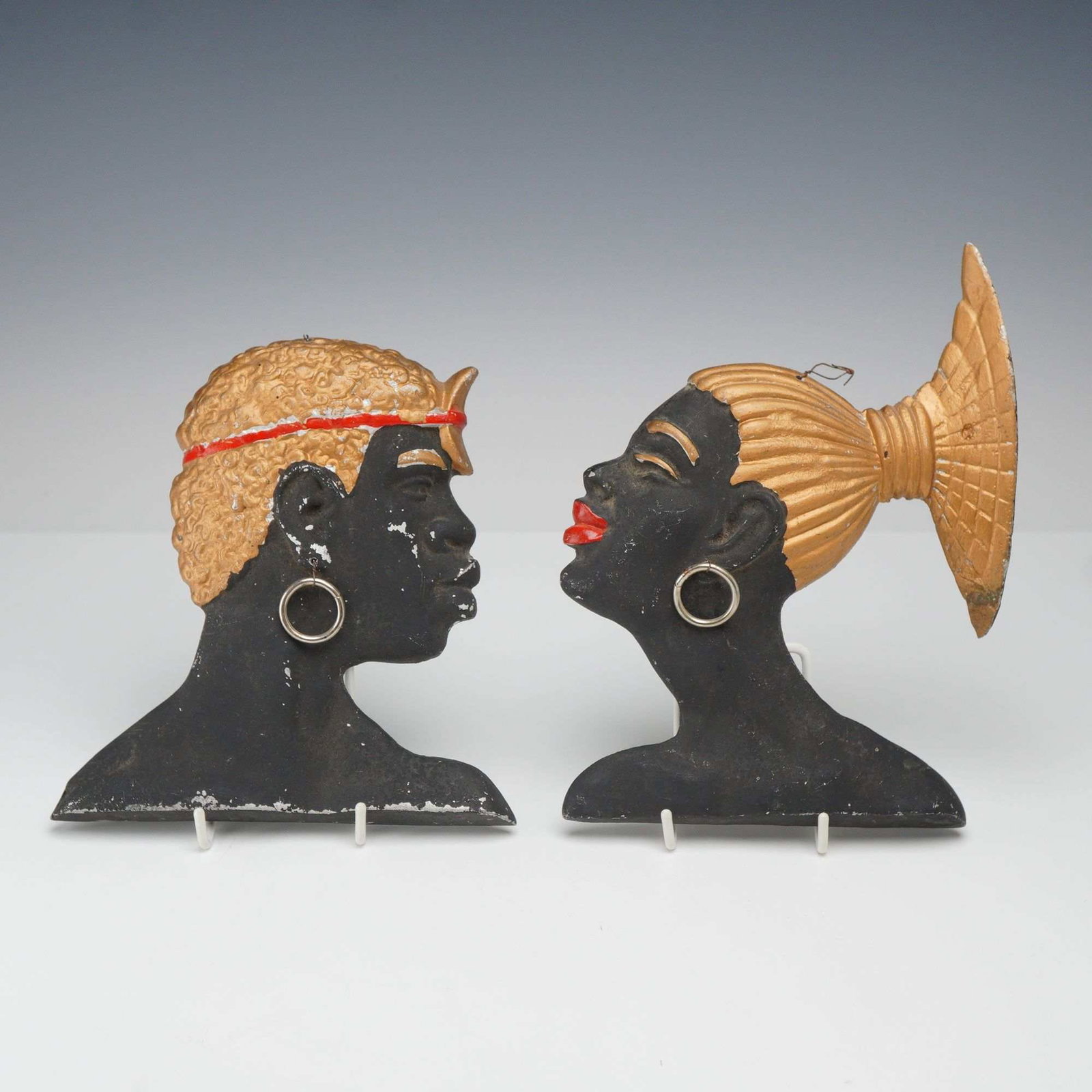 Pair of Mid-Century Modern Painted Cast Aluminum Africana Wall Decorations: A pair of profile wall plaques depicting an African man and woman in a Mid-Century Modern aesthetic. These pieces are constructed from cast aluminum with a cold-painted finish. The male figure is depi