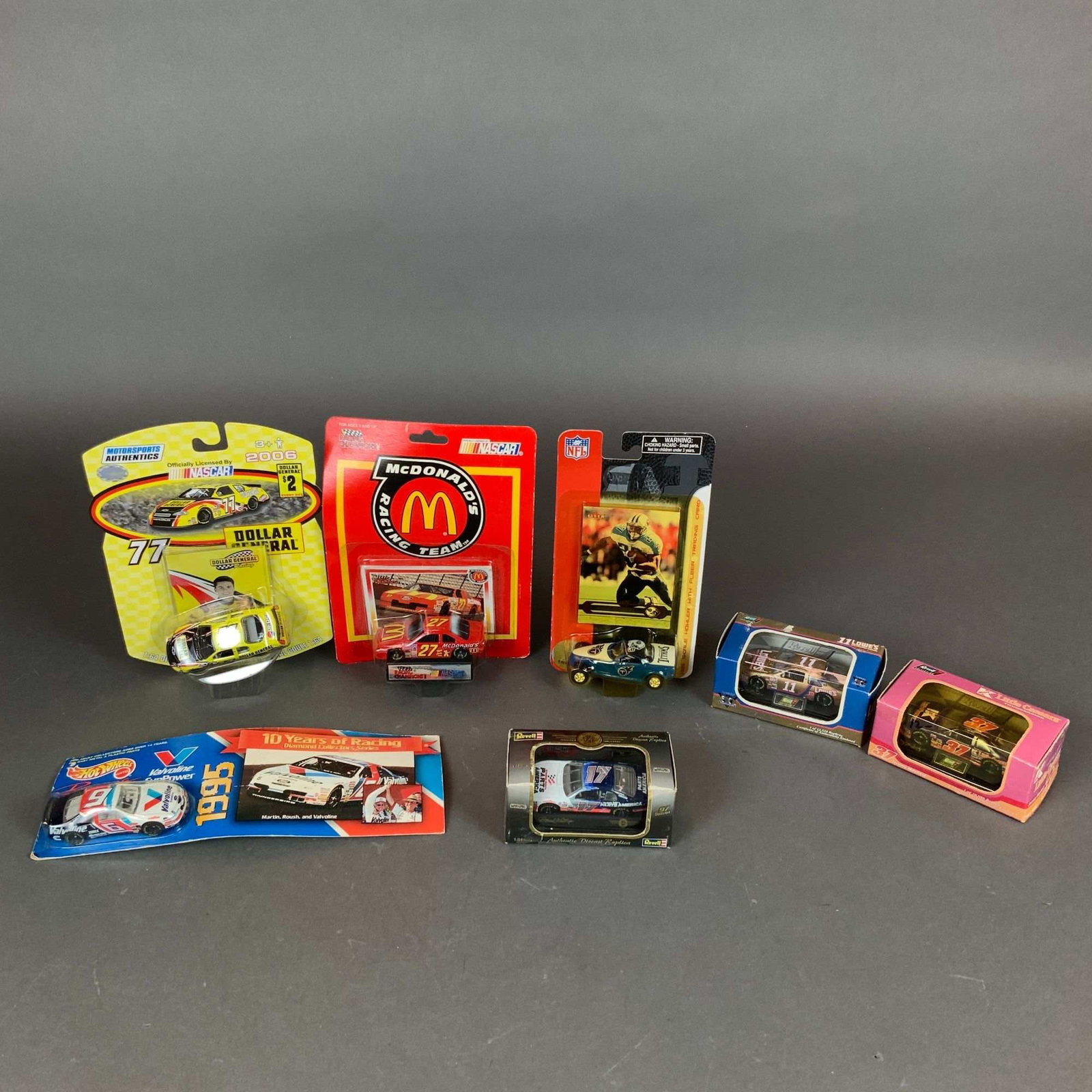 Assortment of Seven NASCAR and NFL Die-Cast Model Cars: This lot consists of seven die-cast collectible vehicles produced by various manufacturers including Motorsports Authentics, Racing Champions, Fleer Collectibles, Hot Wheels, and Revell. The collectio