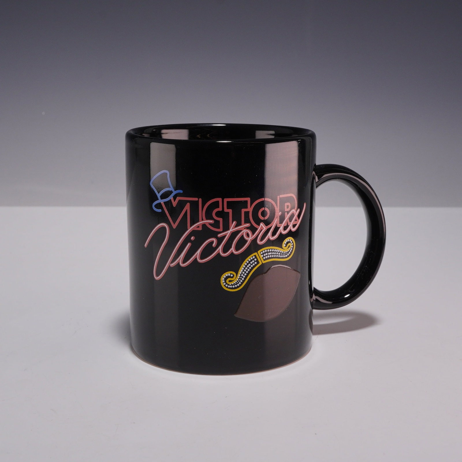 Victor/Victoria Broadway Musical Color Changing Souvenir Mug with Original Care Card: This black ceramic promotional mug was produced for the Broadway musical production of Victor/Victoria. The exterior features a thermochromic neon-style graphic incorporating the show logo, a blue
