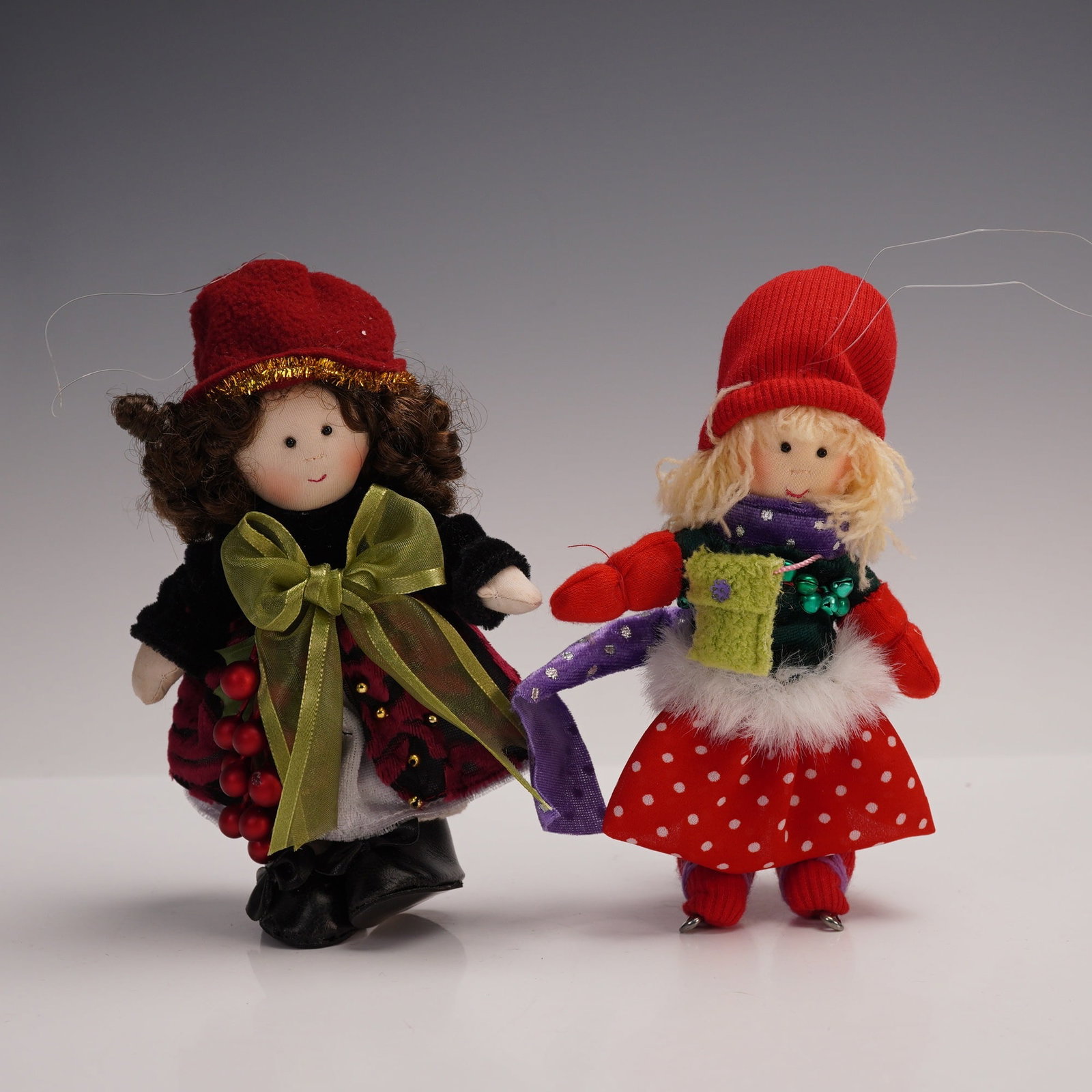 2pc Kurt S. Adler Little Souls Christmas Ornaments: This lot includes two charming fabric dolls, each with distinct holiday attire and accessories. One figure features curly dark hair, a red hat with gold trim, a black velvet-style jacket, a red and bl