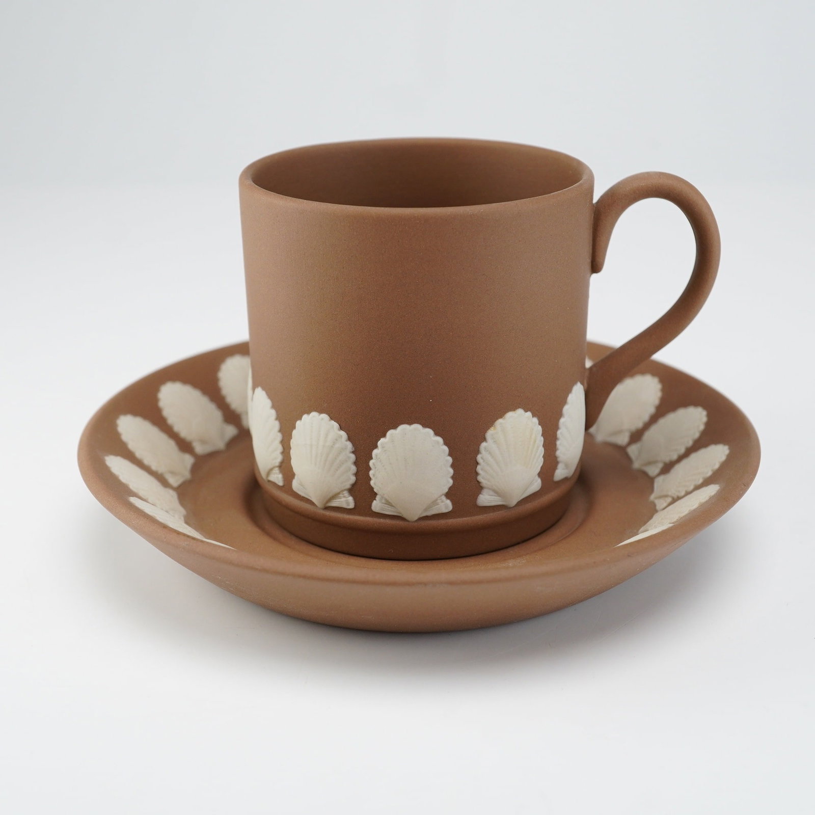 Wedgwood Cream Color Jasperware Sea Shell Demitasse Cup and Saucer Set: A vintage two-piece demitasse cup and saucer set manufactured by Wedgwood in England. The pieces are crafted from unglazed stoneware, specifically taupe or brown jasperware, featuring a matte finish. 