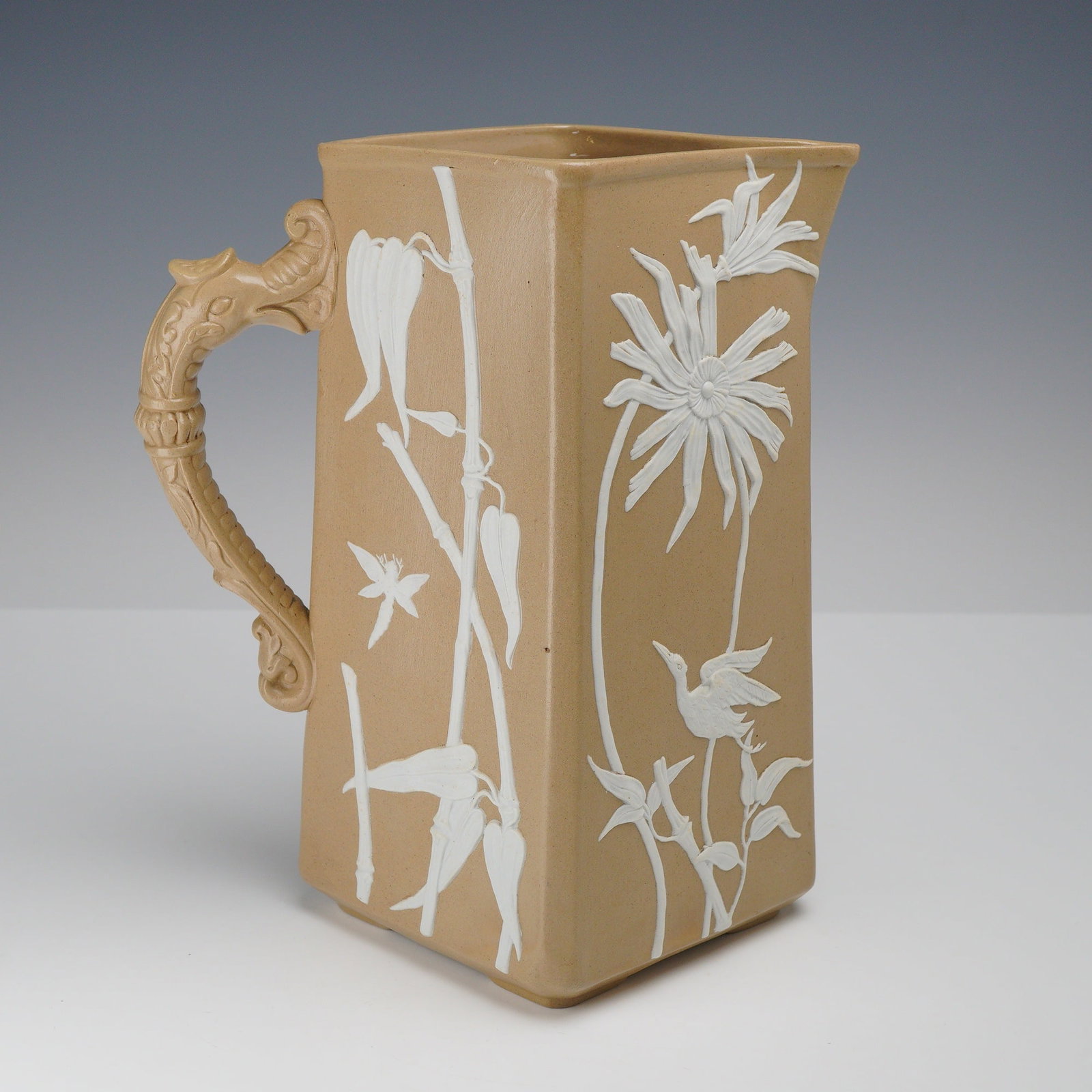 Attributed to Dudson Dragon-Handled Stoneware Pitcher: This rectangular stoneware pitcher, sourced from the Rubin Collection, is attributed to the Dudson pottery of Hanley, Staffordshire. The vessel is crafted from buff-colored stoneware with a matte fini