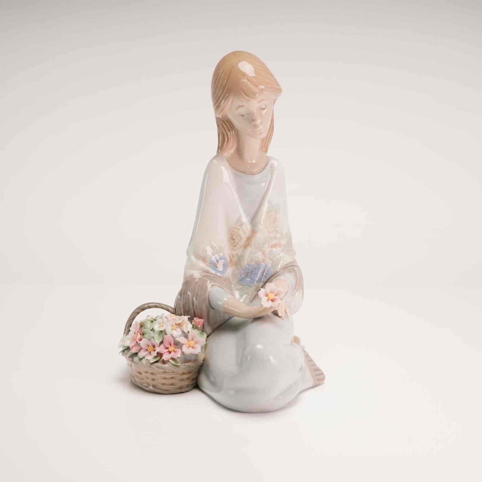 Lladro Porcelain Figurine Flower Song 01007607: This Lladro glazed porcelain figurine depicts a kneeling young woman holding a single blossom. She is dressed in a pale blue gown with a white floral-patterned shawl embellished with cream and blue bl