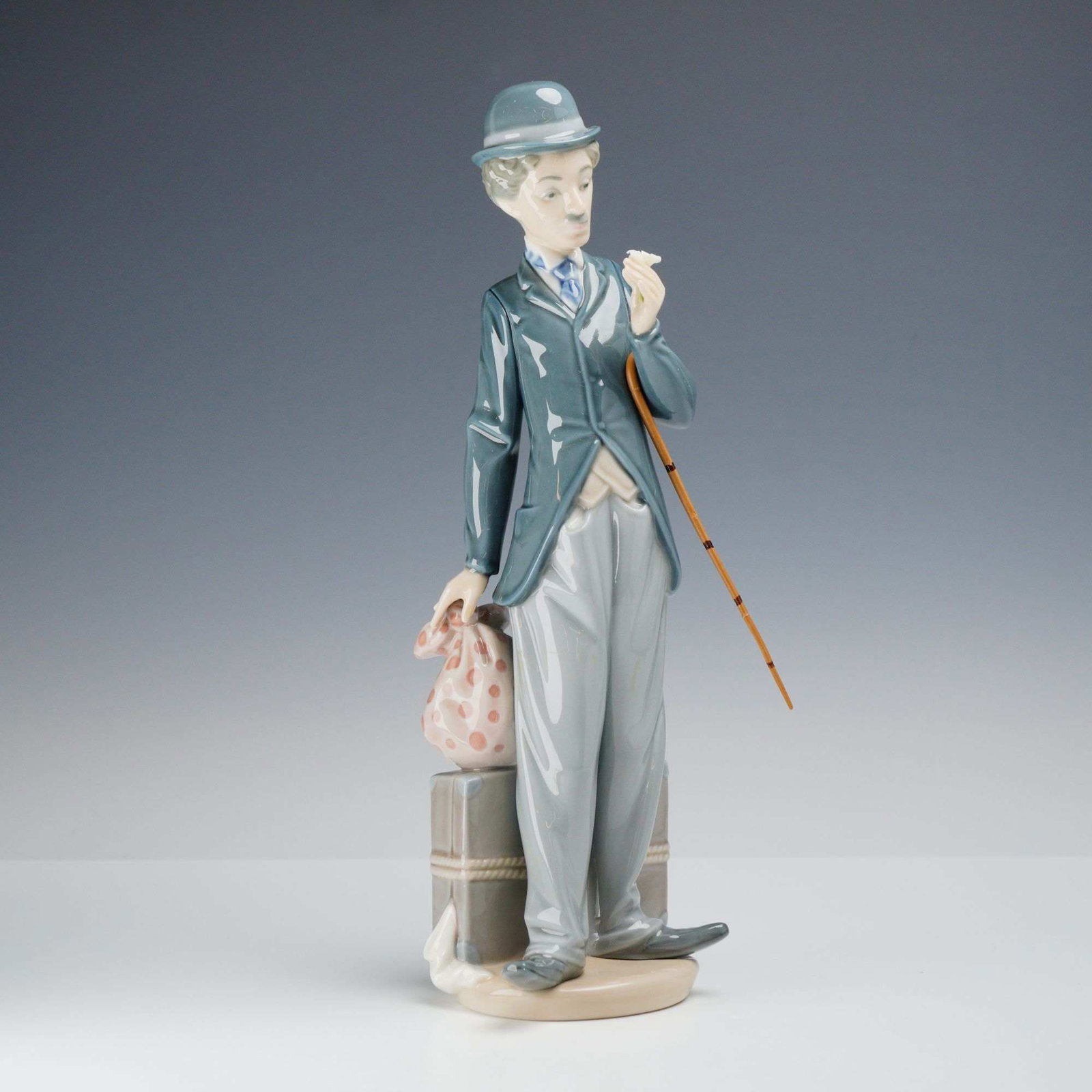 Lladro Porcelain Figurine, Charlie the Tramp 01005233: A glazed porcelain figurine manufactured by Lladro in Spain, depicting the character Charlie the Tramp, item number 01005233. The figure is rendered in a standing pose, leaning against a grey suitcase