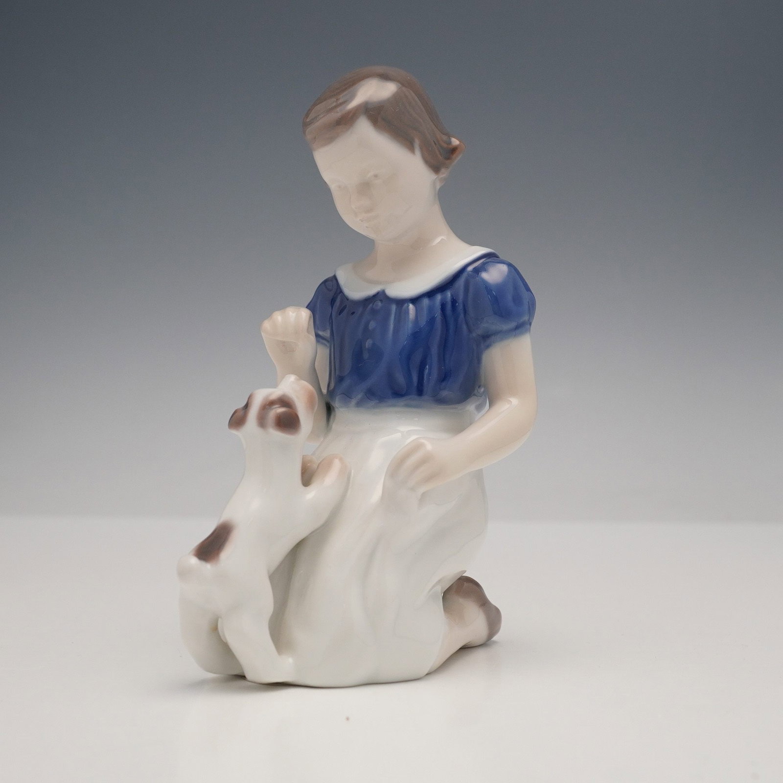 Bing & Grondahl Figurine, Girl With Puppy 2316: This glazed porcelain figurine depicts a young girl kneeling in a blue blouse and white skirt, looking down at and interacting with a small white dog with brown patches. The base bears an underglaze b