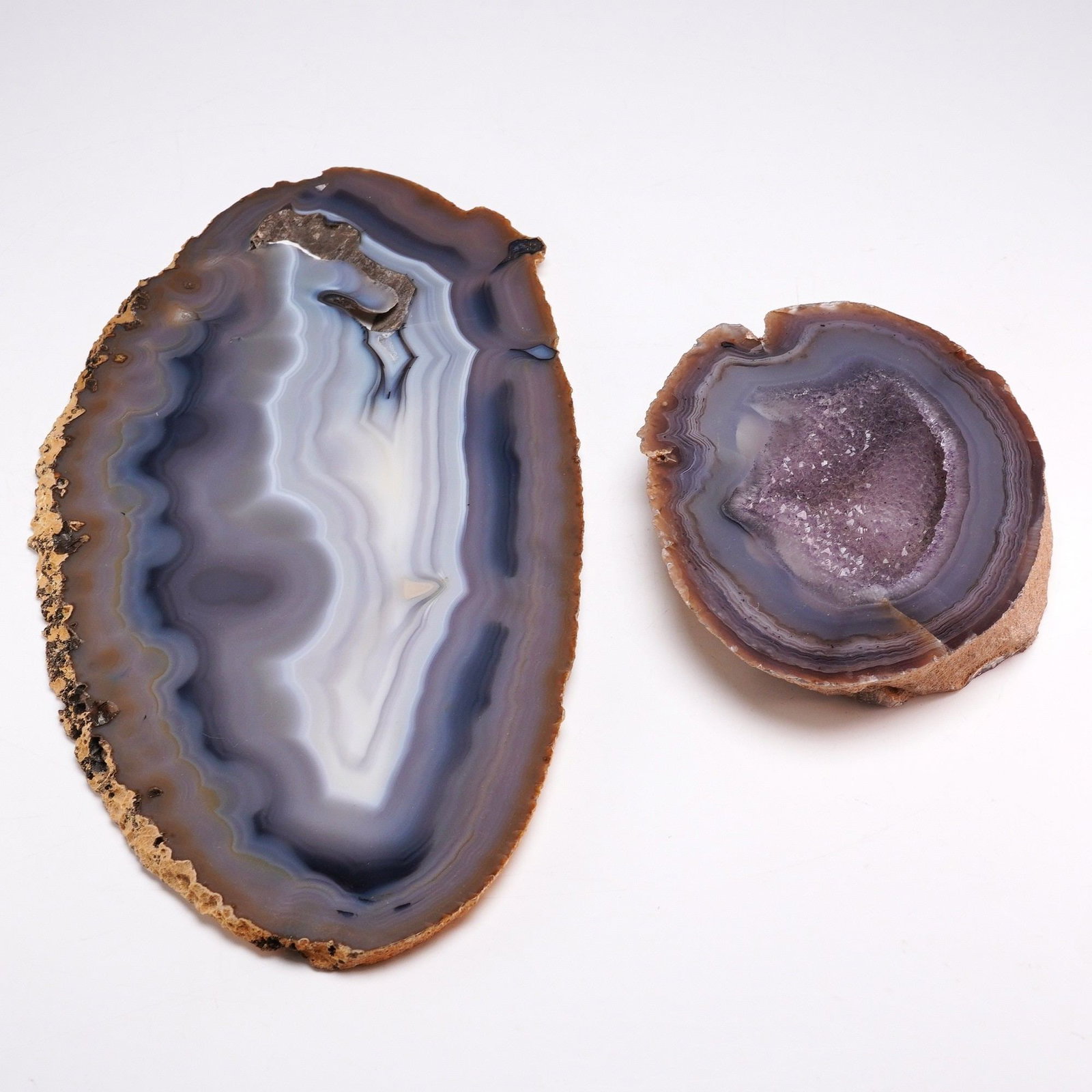 2pc Banded Agate and Geode Slices: A group of two polished mineral specimens, comprising a large oval agate slab and a smaller geode section. The larger slab features concentric banding in variegated shades of gray, white, and
