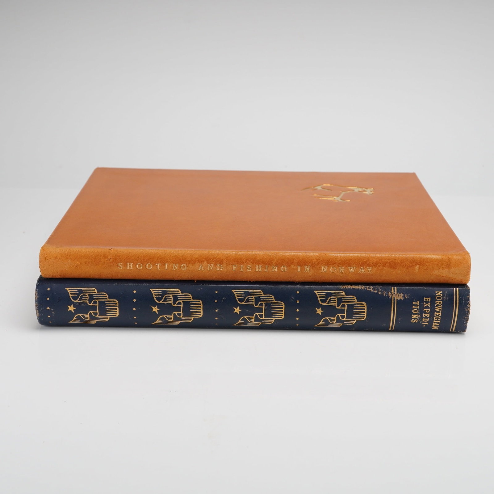 2pc Norwegian Expeditions Hardcover Books: The first volume is bound in orange with the title Shooting & Fishing in Norway lettered in gold on the spine, featuring a small gold pictorial stamp of an animal on the front cover. The second