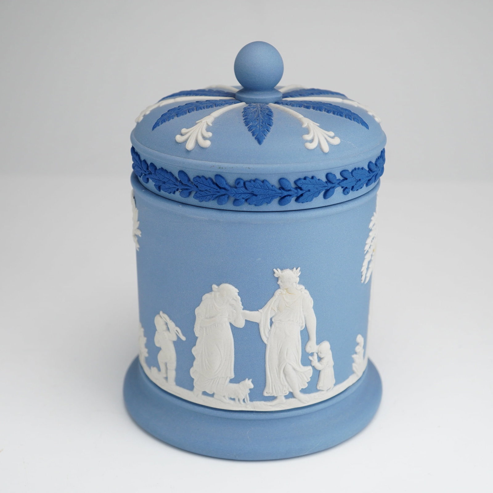 Wedgwood Blue Jasperware Lidded Tobacco Jar: A Wedgwood jasperware lidded jar featuring a cylindrical body on a slightly flared base. The piece is rendered in a pale blue jasper ground and decorated with applied white bas-relief sprigging depict