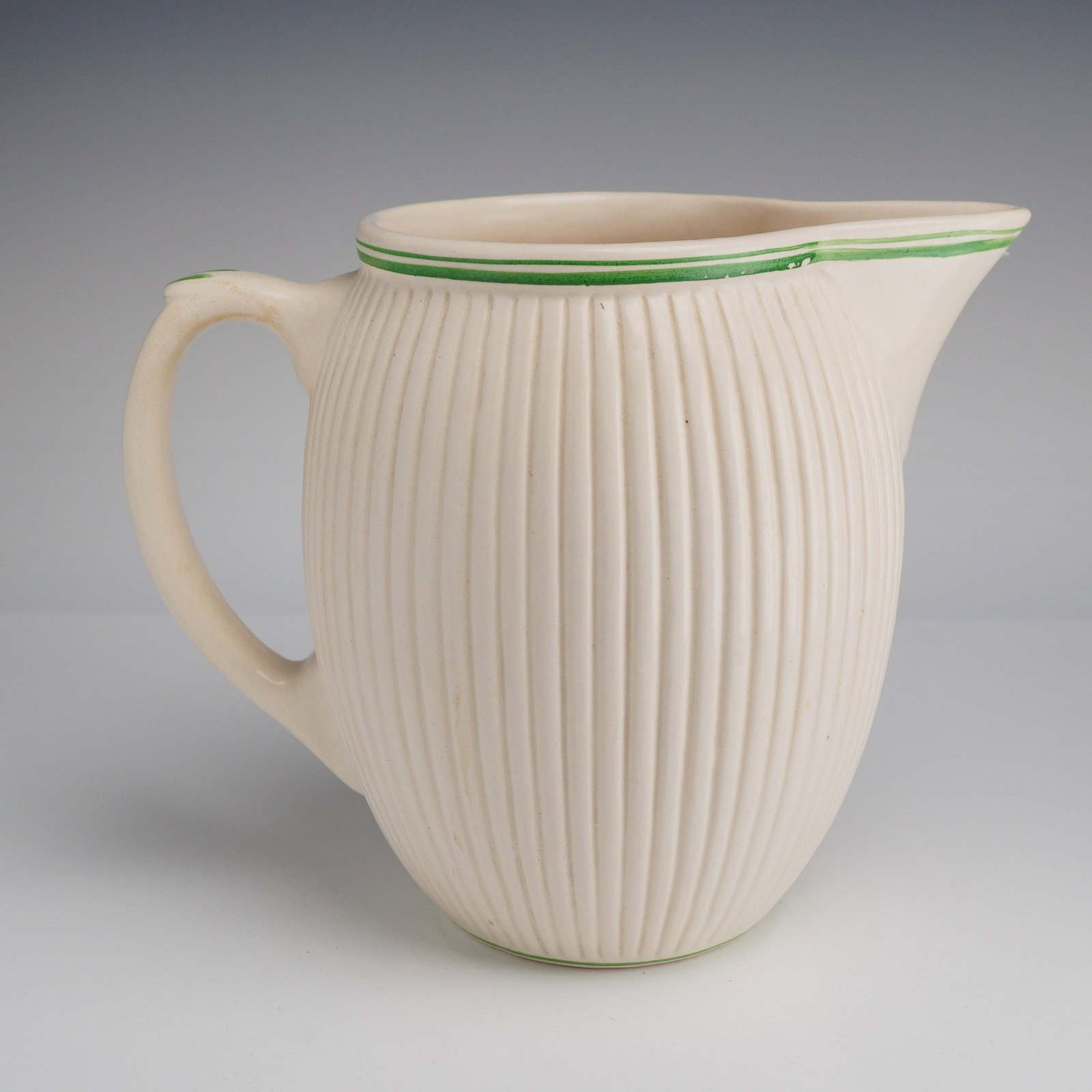 Universal Cambridge Art Pottery Pitcher. Ribbed Design: The vessel features a bulbous, ovoid body decorated with prominent vertical ribbing and finished in an off-white glaze. The rim and base are accented with hand-painted green horizontal bands, with add