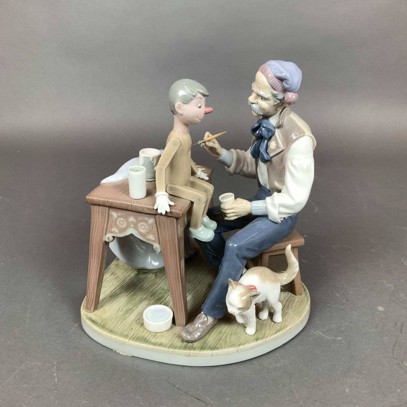 Lladro Glazed Porcelain Figurine, The Puppet Painter 01005396: This glazed porcelain figurine group, titled The Puppet Painter, is produced by Lladro and depicts the character Geppetto from the story of Pinocchio. The scene features Geppetto seated on a stool whi