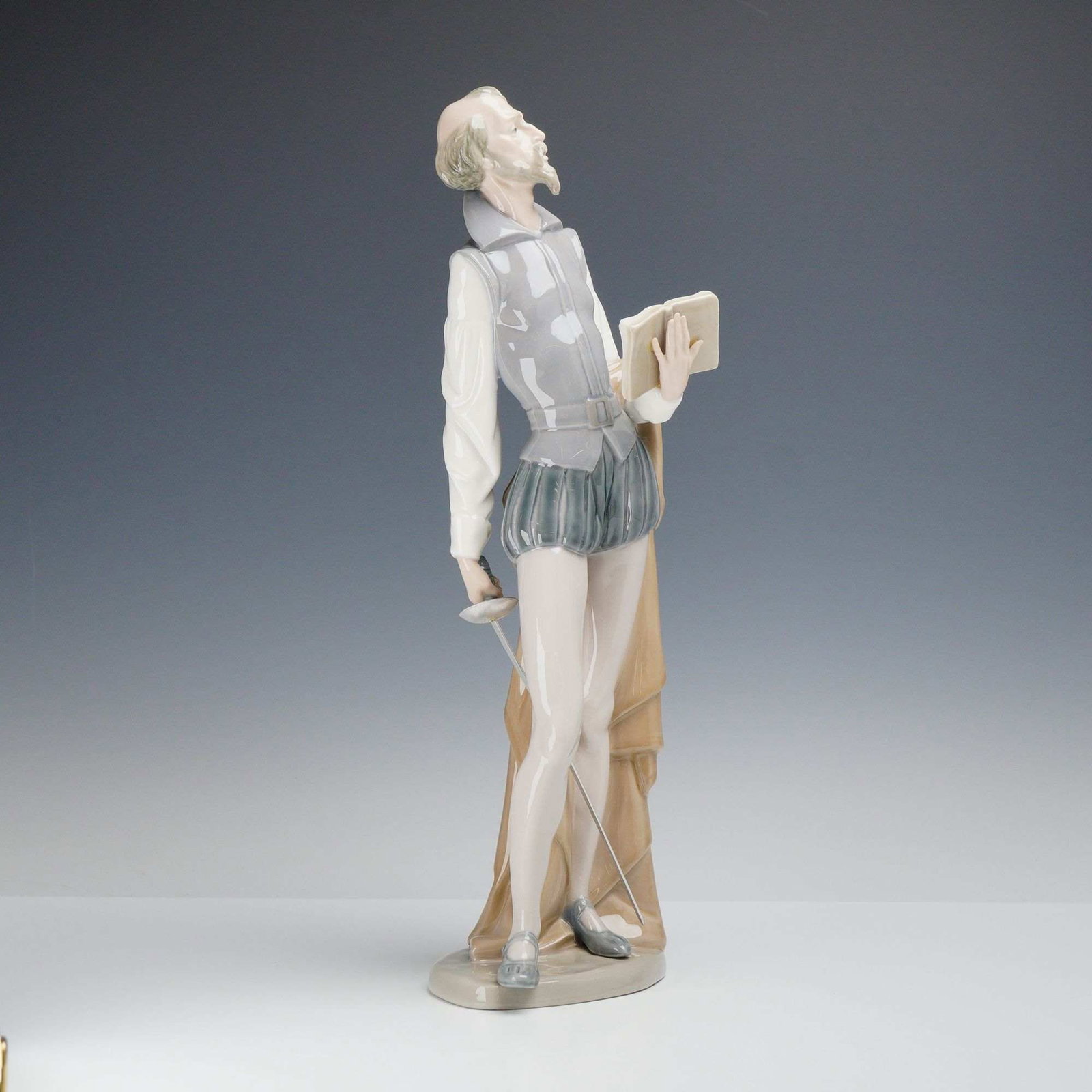 Nao by Lladro Porcelain Figurine, Don Quixote Standing with Book: This glazed porcelain figurine, titled Don Quixote Standing with Book, represents the character Don Quixote Reading with Sword as produced in the Nao by Lladro style. The figure is rendered in an elon