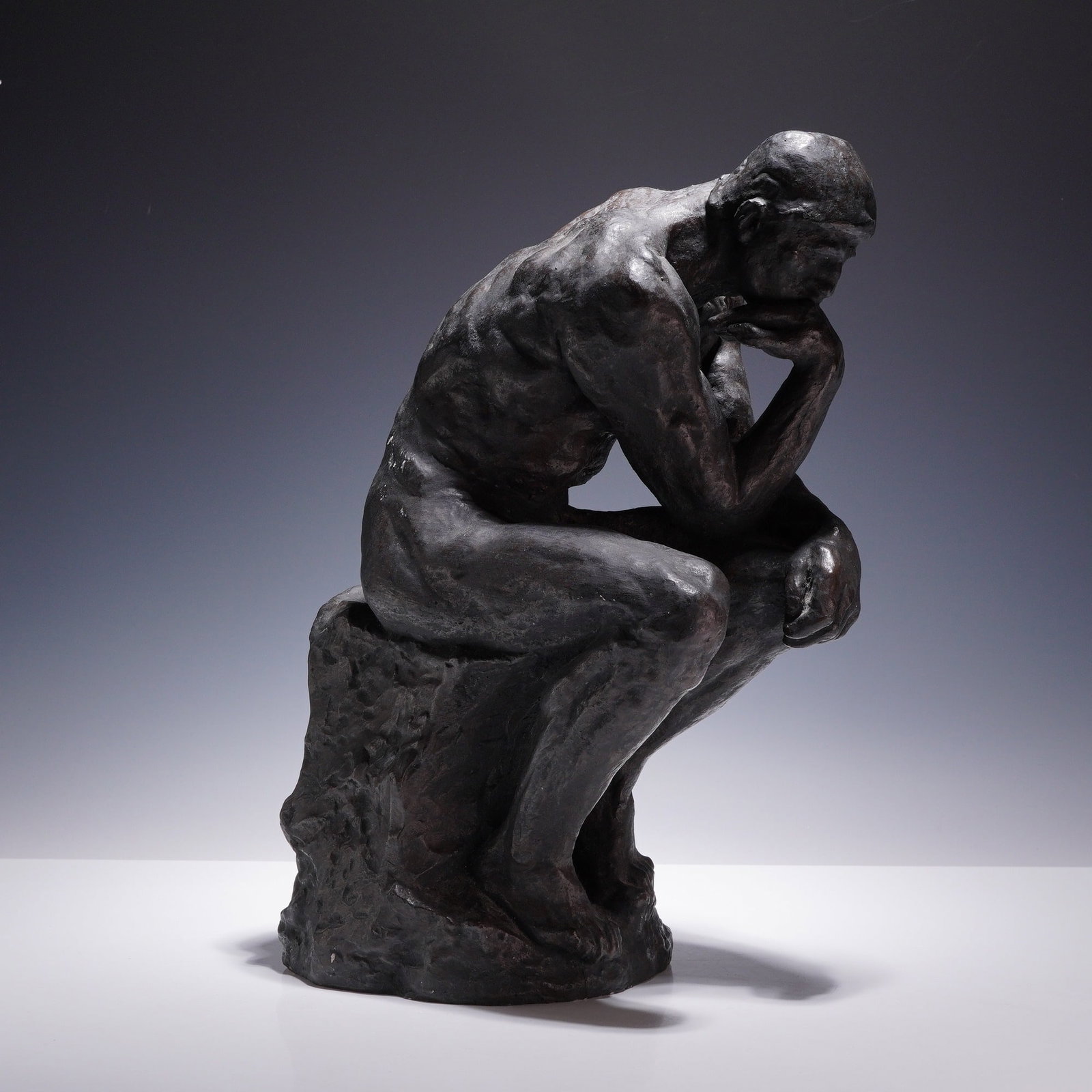 After Auguste Rodin, The Thinker, a Metal Figure: This sculpture is a reproduction of Auguste Rodin's iconic work, The Thinker, rendered as a metal figure. The piece depicts a muscular nude male figure in a seated, contemplative pose with his chin re