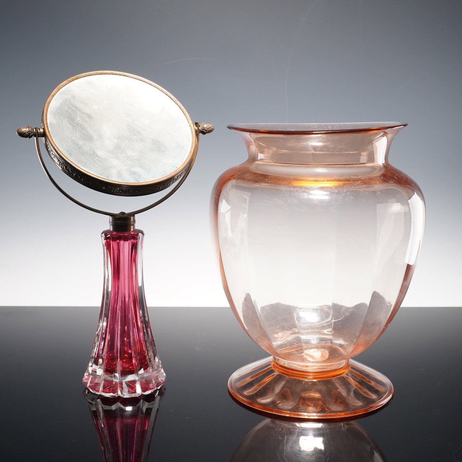 2pc Vanity Mirror and Pink Vase: Group of two glass items including a vanity mirror with ruby-to-clear cut glass base and metal frame with embossed floral detail, together with a pink pressed glass vase of rounded form on a flared
