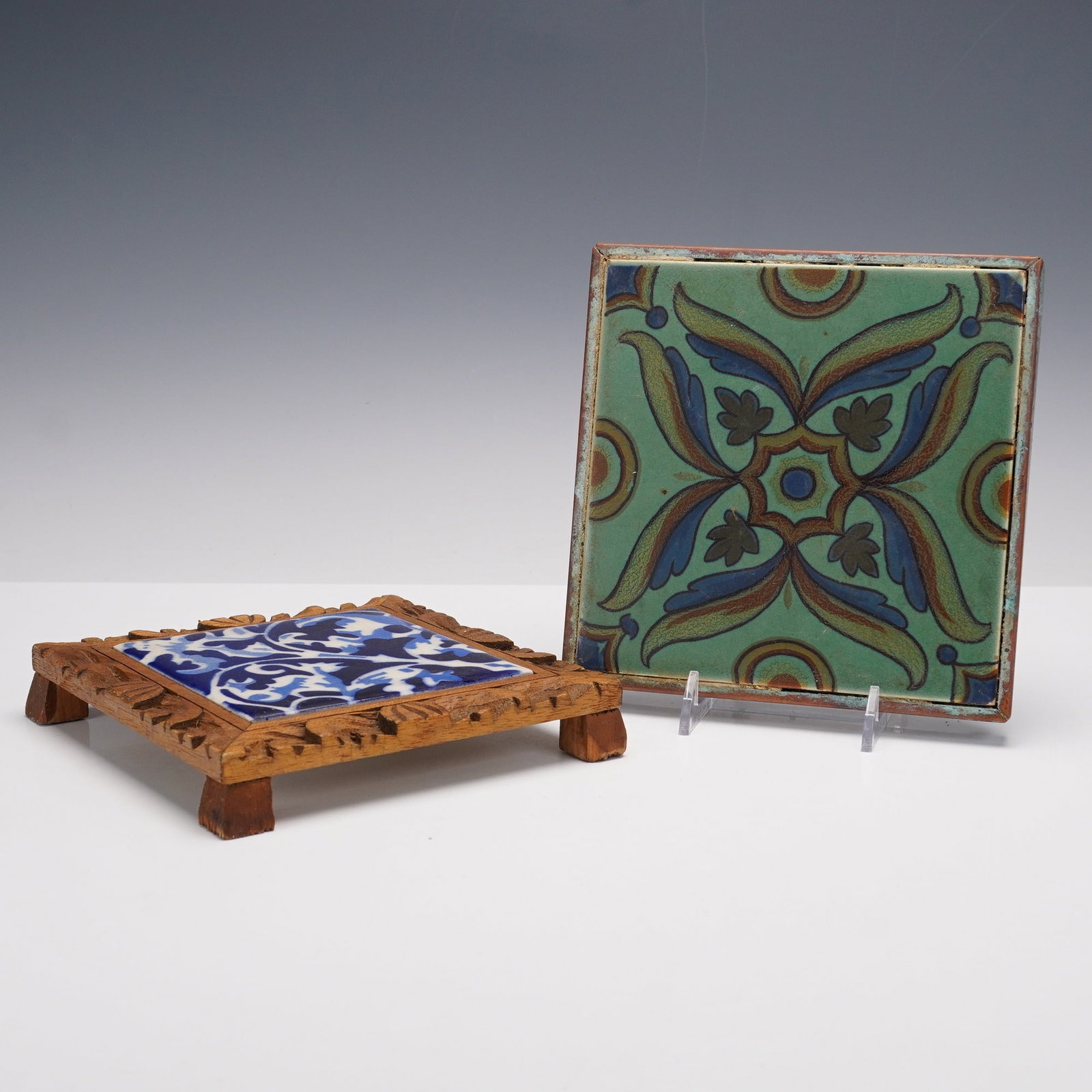2pc Vintage Decorative Tiles Trivets: A lot of two vintage decorative tiles including a square green ground tile with stylized foliate pattern in blue and brown tones displayed on a stand, together with a blue and white foliate patterned