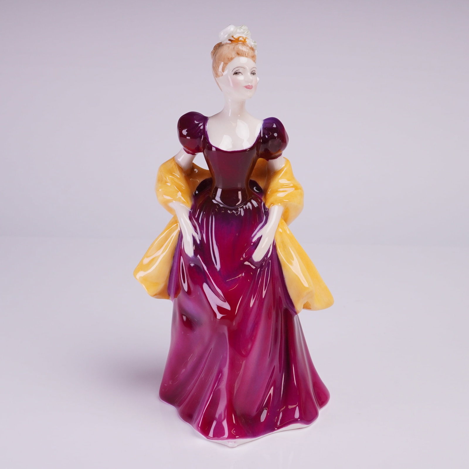 Royal Doulton Figurine Loretta HN2337: The figure depicts a standing woman in a period-style formal gown, featuring a deep magenta and purple polychrome glaze on the skirt and bodice, complemented by a bright yellow shawl draped over the s