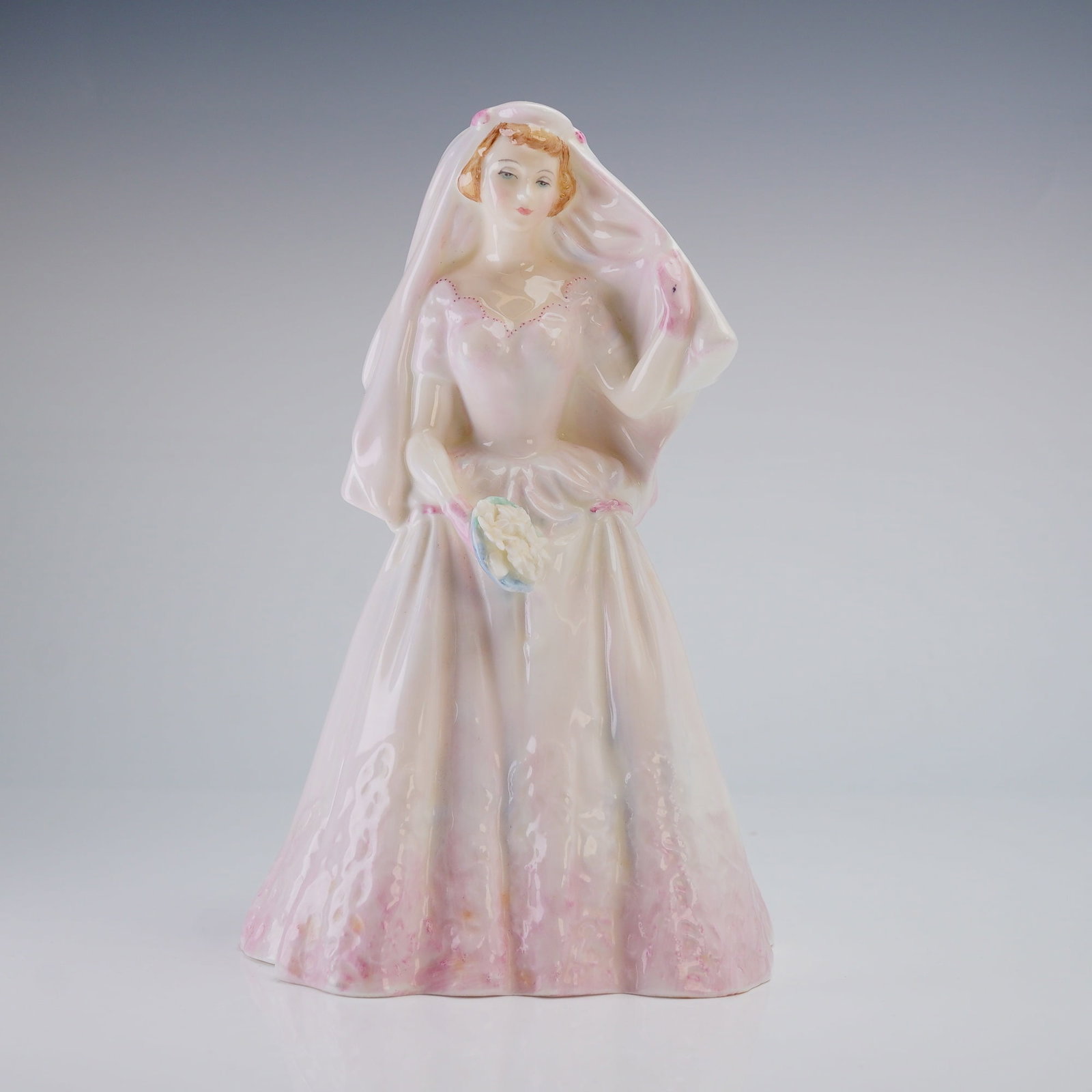 Royal Doulton Figurine The Bride HN2166: This mid-20th-century piece features a woman in a wedding gown with a pale pink wash and deeper pink accents at the hemline. The figure is depicted wearing a long veil and carrying a small bouquet. Th