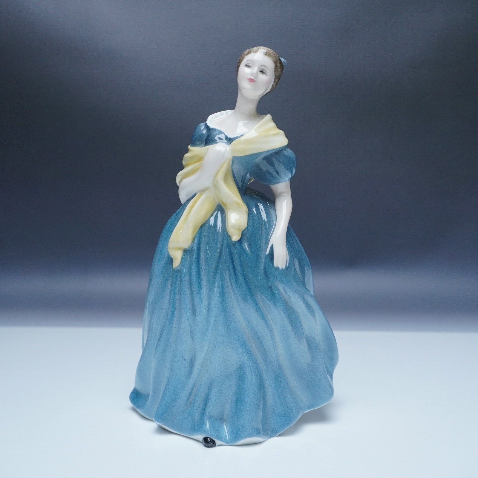 Royal Doulton Figurine, Adrienne HN2304: This Royal Doulton figurine, named "Adrienne" (HN2304), stands at 8"H. Designed by Peggy Davies, it was issued between 1964 and 1991. The figurine portrays a lady in a teal blue dress with a yellow sh