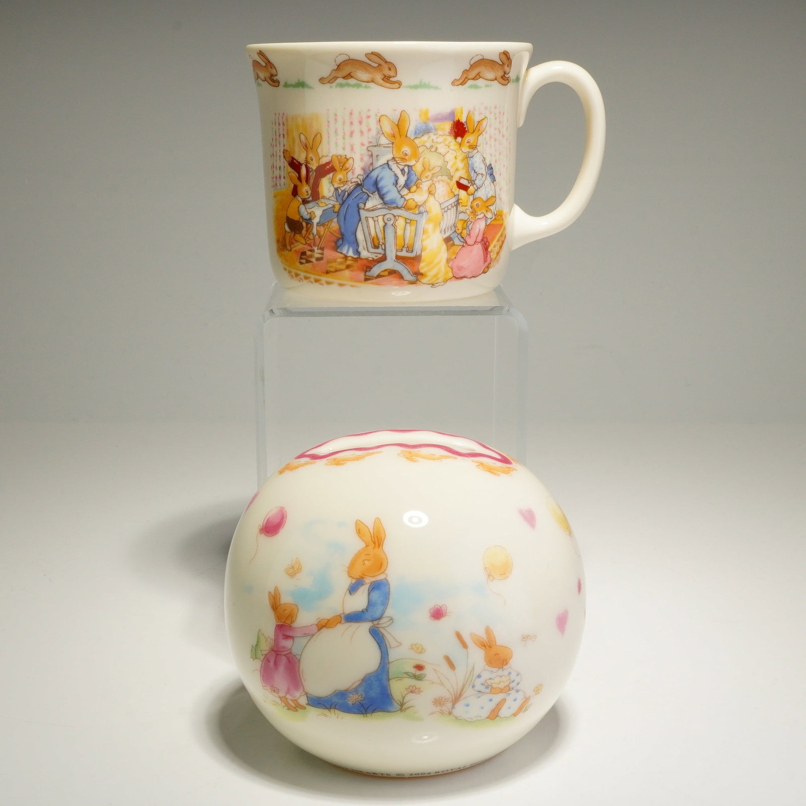 2pc Royal Doulton Bunnykins Christening Mug and Sweet Hearts Money Bank: This collection features a handled mug and a spherical money bank, both crafted from bone china with a cream-colored ground. The mug is decorated with transfer-printed illustrations of rabbit