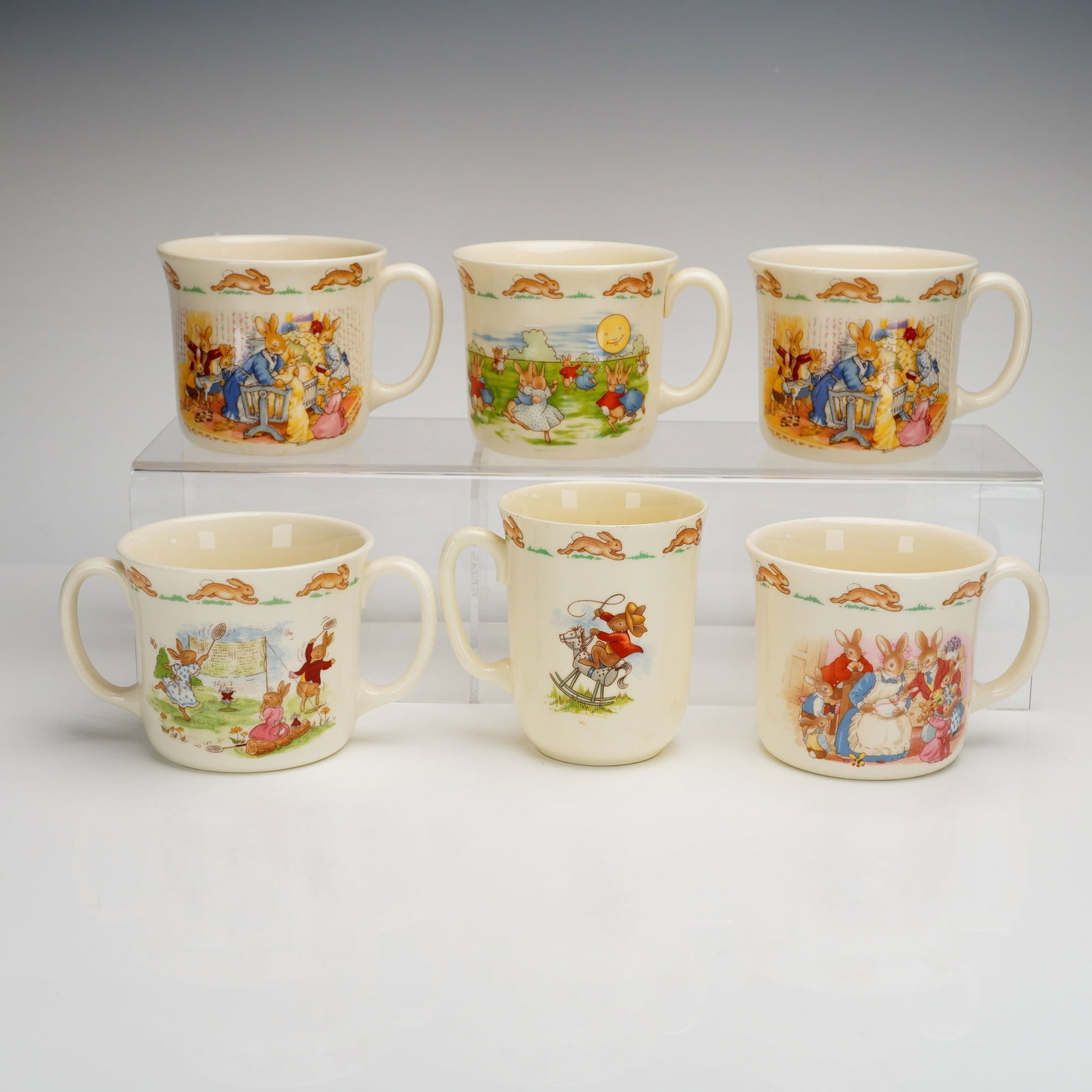 6pc Royal Doulton Bunnykins Bone China Mugs & Two-Handled Cup: A collection of six Royal Doulton Bunnykins fine bone china child's mugs and a two-handled cup, also known as a hug-a-mug. The items feature classic transfer-printed illustrations of anthropomorphic