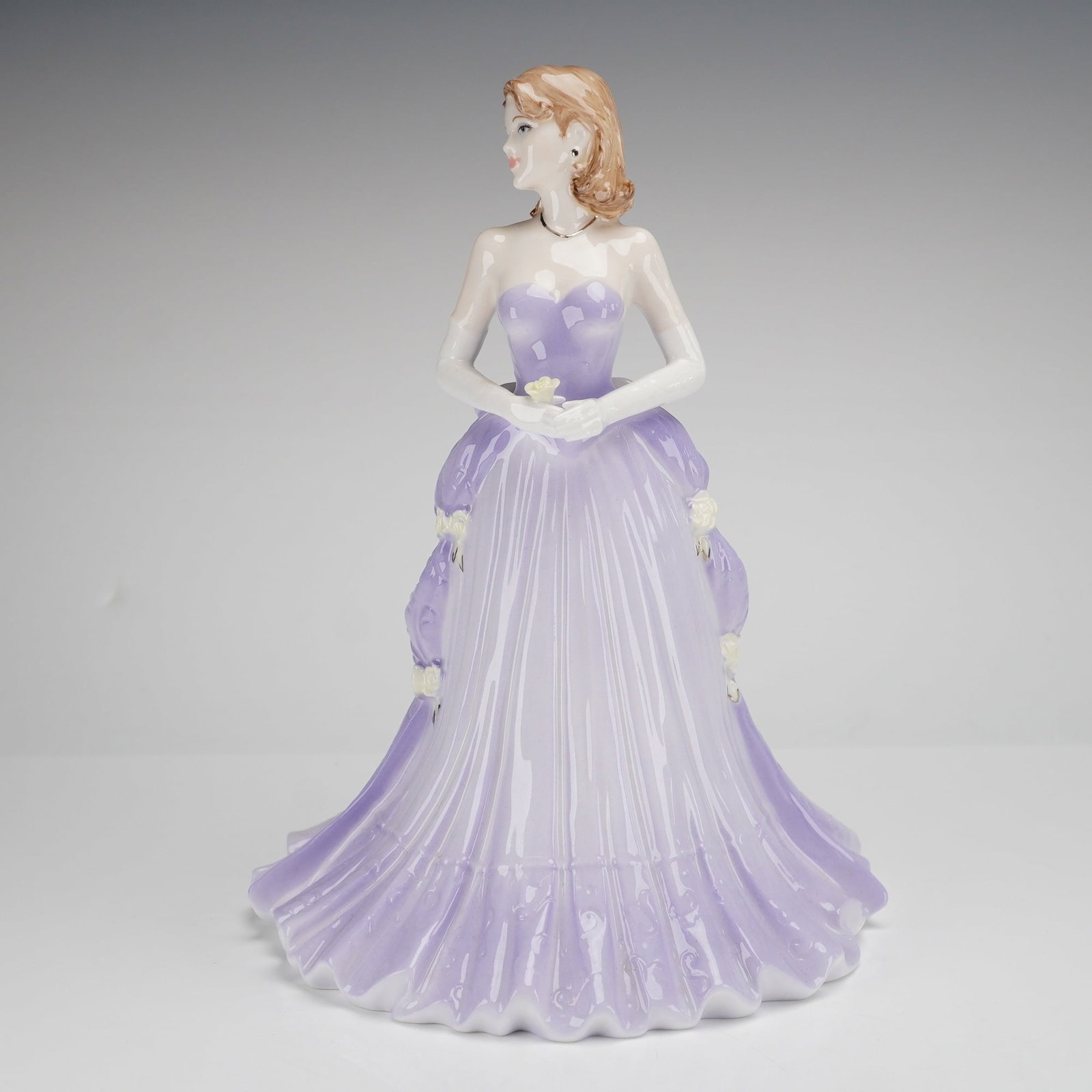 Coalport Bone China Figurine, 2005 May Ball: The figure is constructed of glazed bone china and depicts a standing female figure with light brown hair. She is dressed in a lavender-colored evening gown featuring a strapless, corset-backed