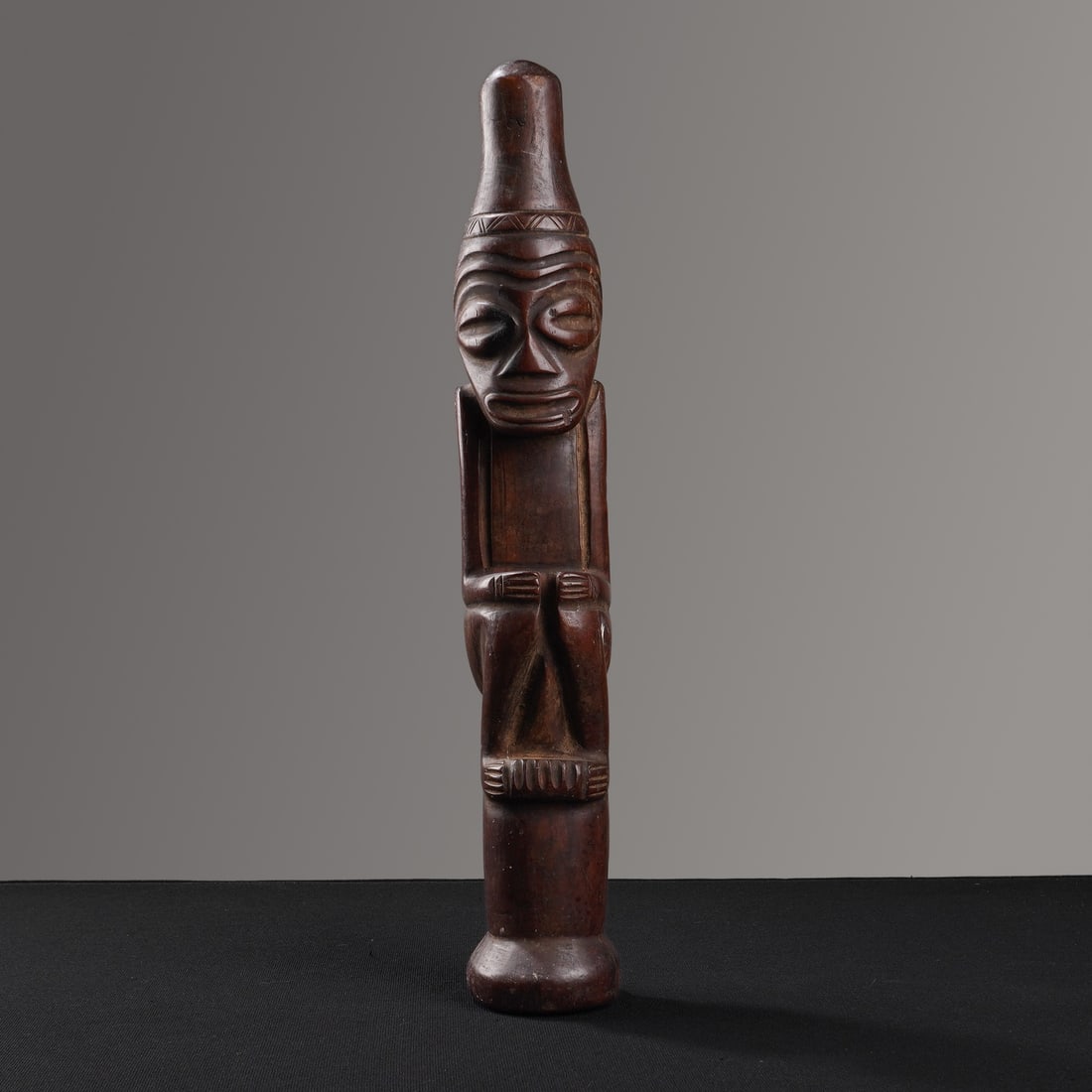 African Wood Carved Seated Figure: This wood carved sculpture depicts a stylized human form in a seated posture. The figure is hand-carved from a single block of dark-stained solid wood and is positioned atop a cylindrical pedestal wit