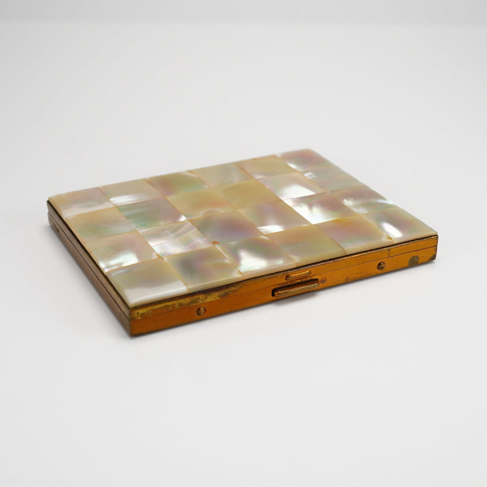 Mother of Pearl and Brass Compact Case: This rectangular compact case features a decorative checkerboard inlay of iridescent mother-of-pearl tiles on the front cover, set within a brass frame. The reverse of the case consists of a plain bra