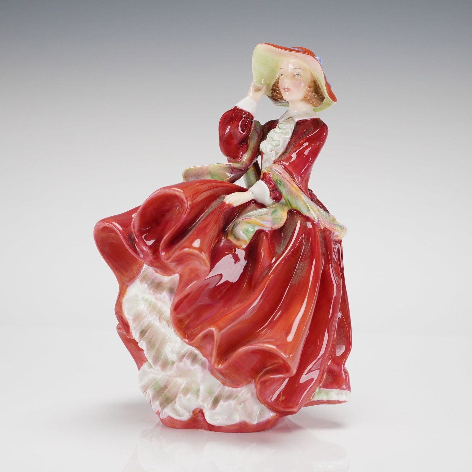 Royal Doulton Figurine Top of the Hill HN1834: A single glazed porcelain figurine by Royal Doulton, titled Top of the Hill, model number HN1834. The figurine depicts a lady in a voluminous, high-gloss crimson red gown, posed as if bracing against 
