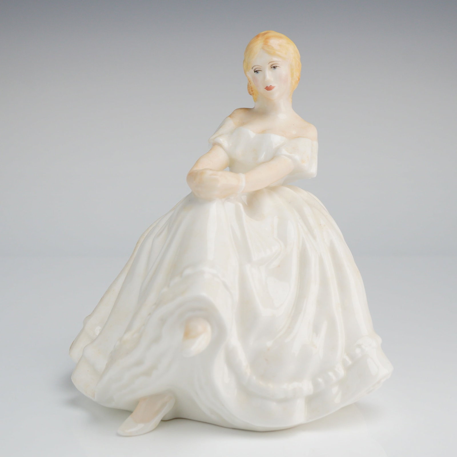 Royal Doulton Figurine Heather HN2956: The figure depicts a seated young woman wearing a glazed, off-the-shoulder white dress in a Romantic or Victorian style. The figure is hand-painted with light details on the face, hair, and bodice. Th