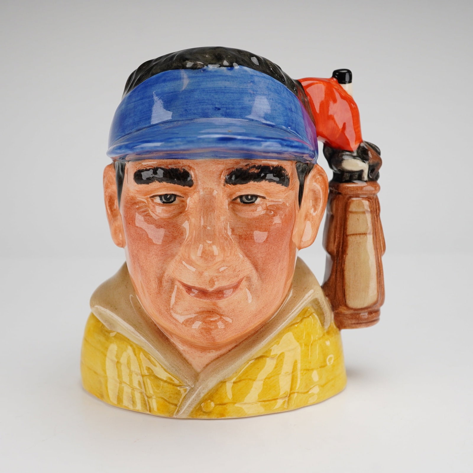 Royal Doulton Small Character Jug, The Golfer D7064: This piece was modeled by David B. Bigg and carries a copyright date of 1997. The figure is depicted with a fair complexion, wearing a blue sun visor and a yellow collared shirt. The handle is designe
