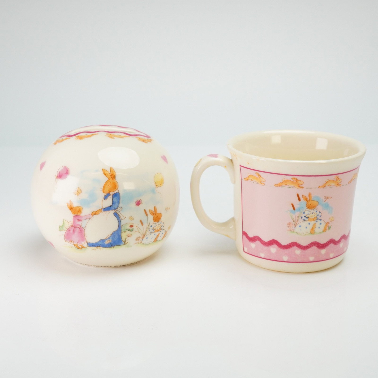 2pc Royal Doulton Bunnykins Bone China Money Ball and Mug: This set of Royal Doulton Bunnykins nursery ware is presented in the Sweetheart pattern, identified by the reference H5317. The collection comprises a spherical bone china money ball and a single-hand
