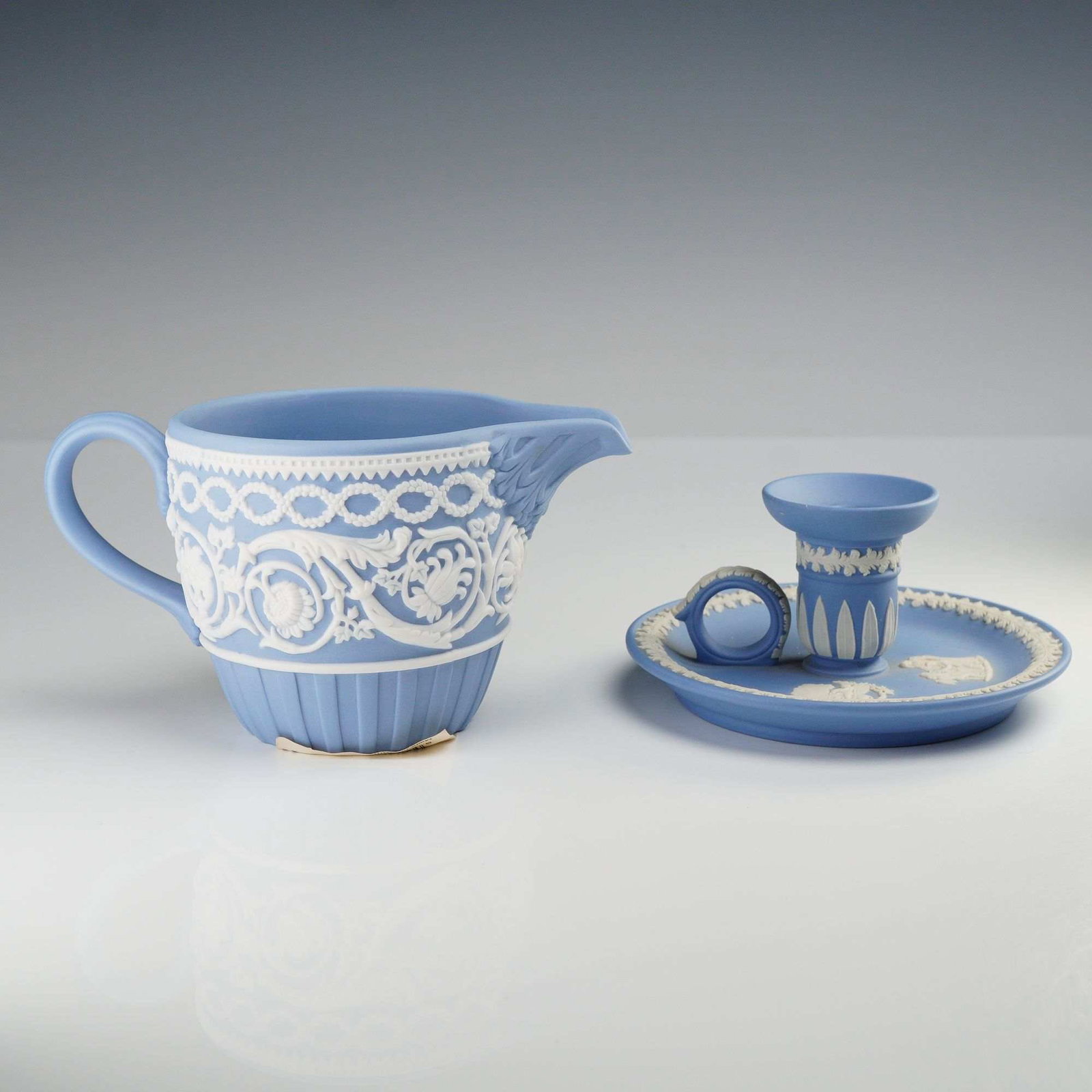 Two Wedgwood Pale Blue Jasperware Pieces: 250th Anniversary Creamer and Chamberstick: This lot consists of two pieces of Wedgwood pale blue jasperware, a signature unglazed matte stoneware featuring white bas-relief sprigged decoration. The first item is a commemorative creamer or
