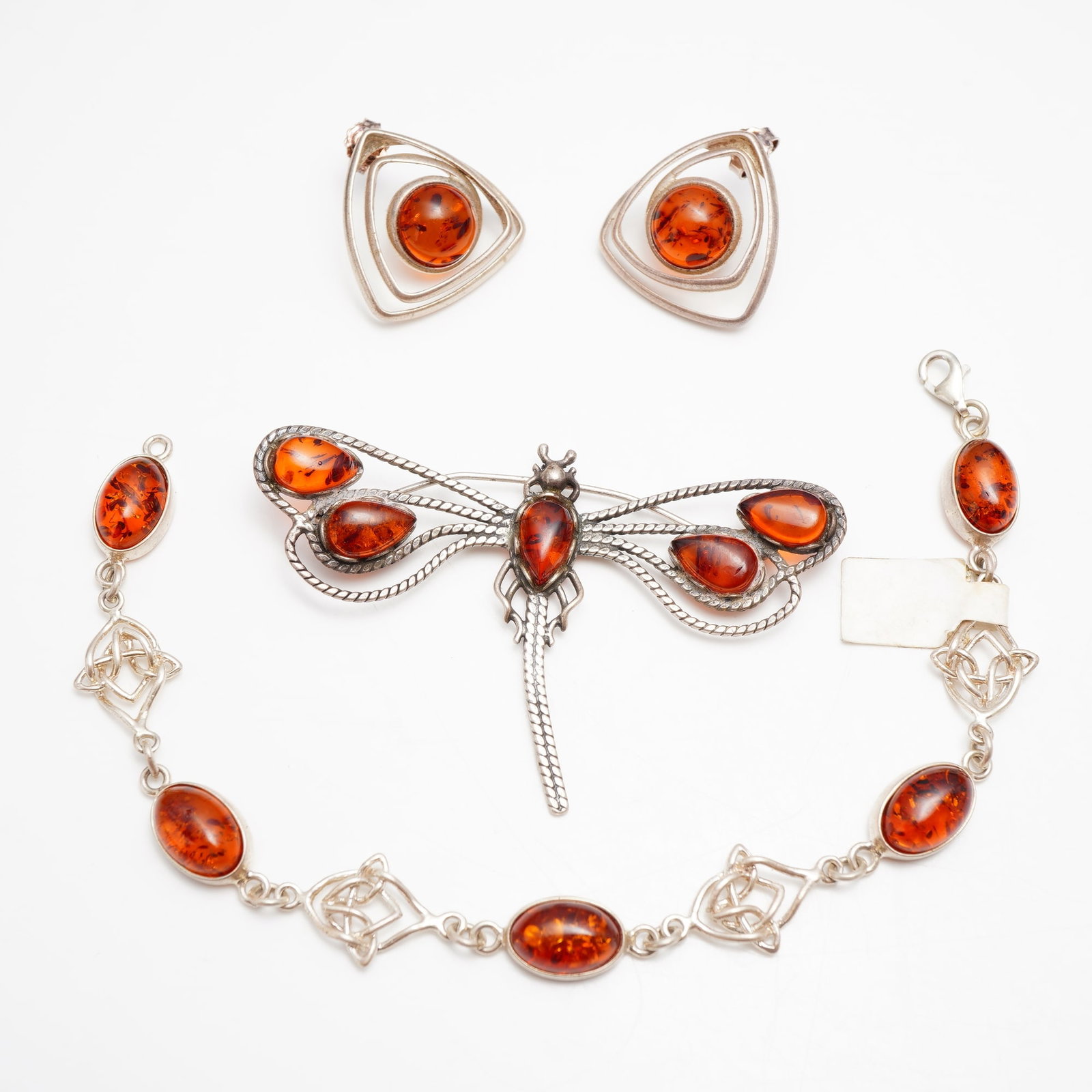 Collection of Four Sterling Silver and Amber Jewelry- Dragonfly Brooch, Celtic Knot Bracelet, and: This set of cognac Baltic amber and sterling silver jewelry includes one pin, one link bracelet, and one pair of stud earrings. The dragonfly brooch is designed with a textured, segmented body and fiv