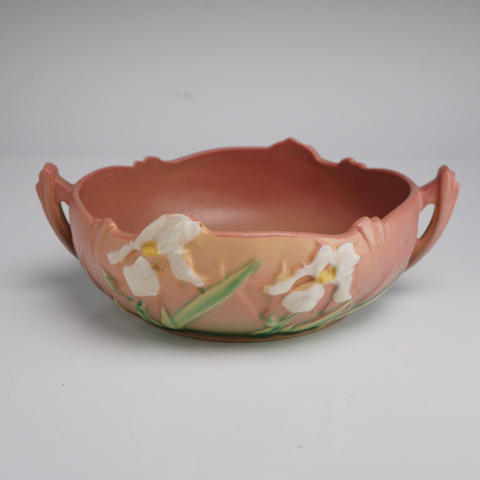 Roseville Pottery Console Bowl, Iris: This ceramic vessel features relief-molded white iris blossoms with yellow centers and green foliage set against a matte, dusty rose-colored ground. The form is characterized by a low profile, a scall