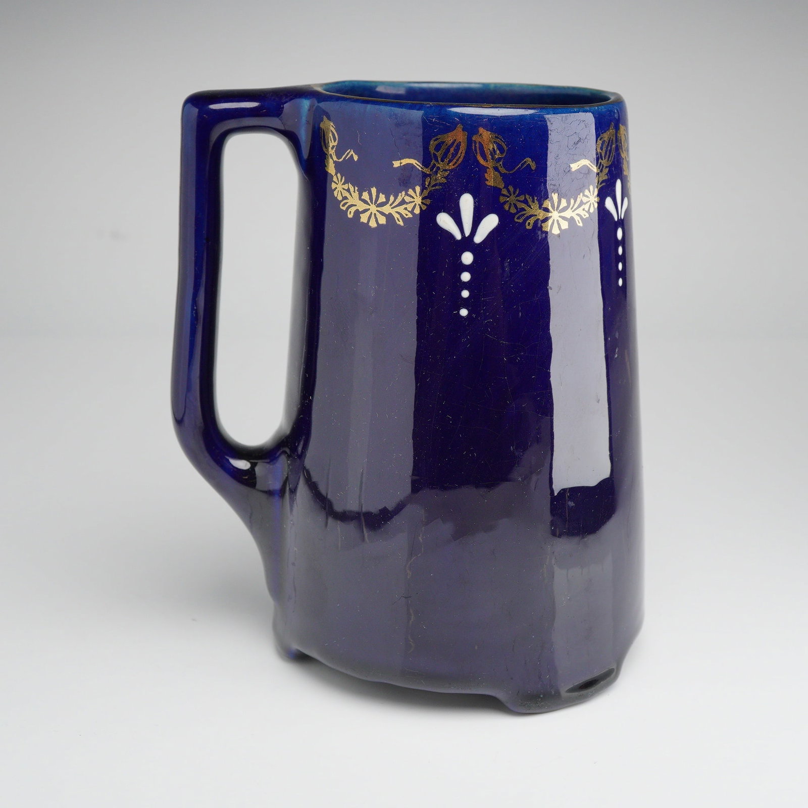 Rare Early Roseville Pottery Jug with Sprayed Gold Accents: This cobalt blue jug features a deep high-gloss glaze over a tall, slightly tapered body. The exterior is decorated with sprayed gold accents in a floral swag pattern near the rim, interspersed with h