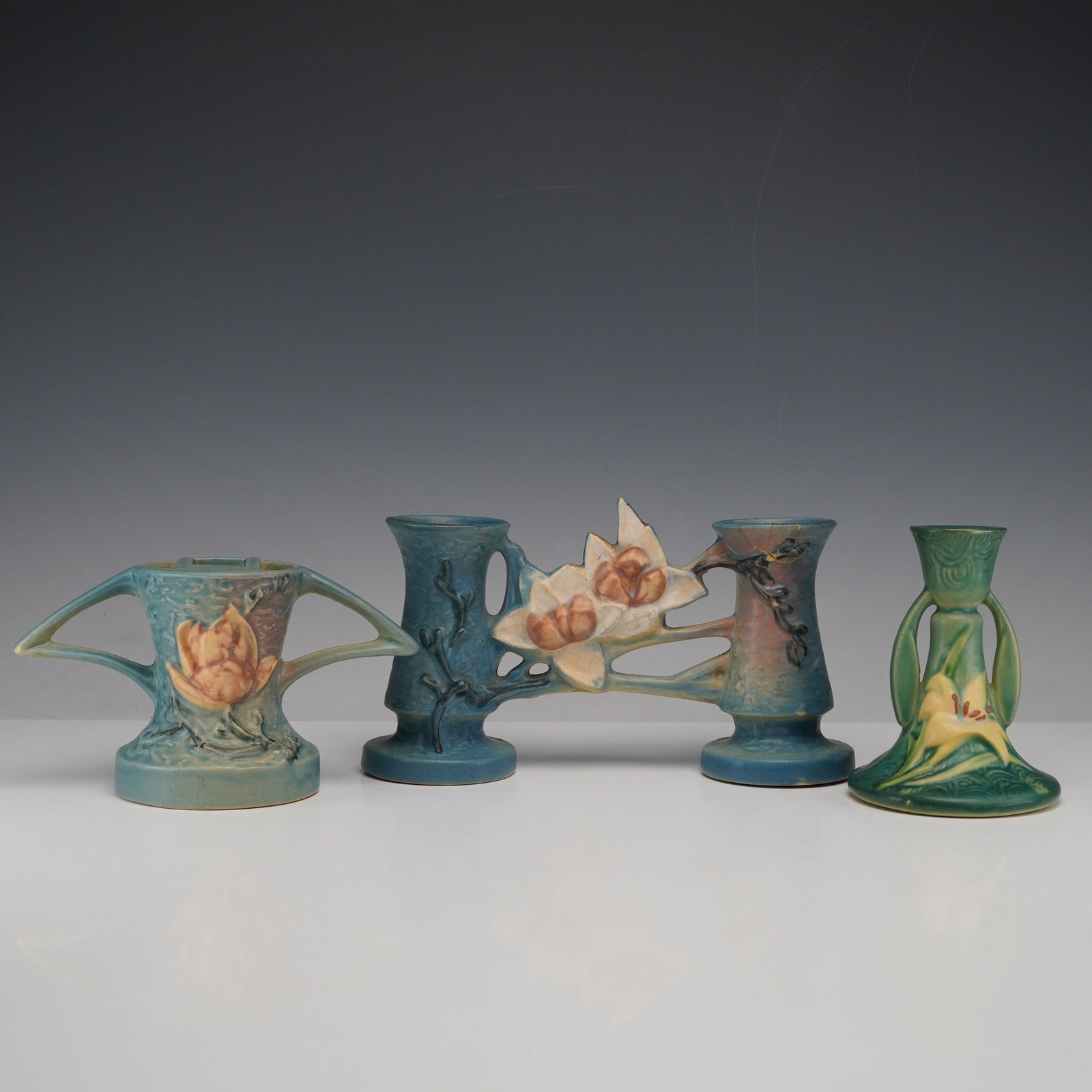 3pc Roseville Pottery Flower Frog, Vase, and Candlestick: Three-piece group of Roseville pieces comprising a Magnolia flower frog and double bud vase, both in blue, and a Zephyr Lily candlestick in green. Each shows molded floral decoration with matte glazes