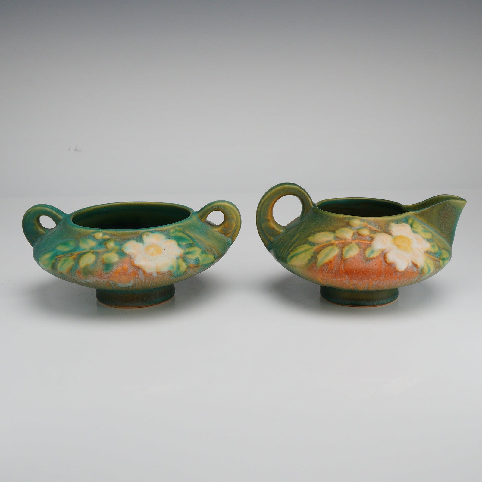 Roseville Pottery Creamer and Sugar Bowl Set, White Rose: A two-piece American art pottery set manufactured by the Roseville Pottery Company of Zanesville, Ohio, in the White Rose pattern, introduced circa 1940. The set comprises a creamer and an open sugar 