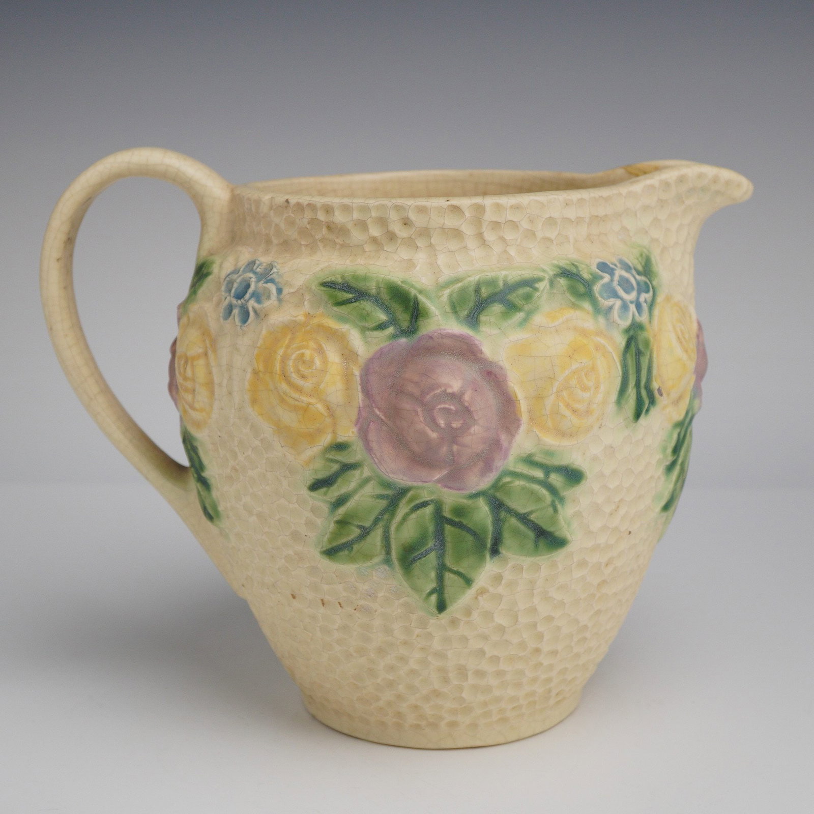 Roseville Pottery Ceramic Pitcher, Rozane 1917: This item features a relief-molded floral band of roses and small blossoms in shades of pink, yellow, blue, and green. The bulbous body is finished in a cream-colored glaze with a hammered or dimpled 