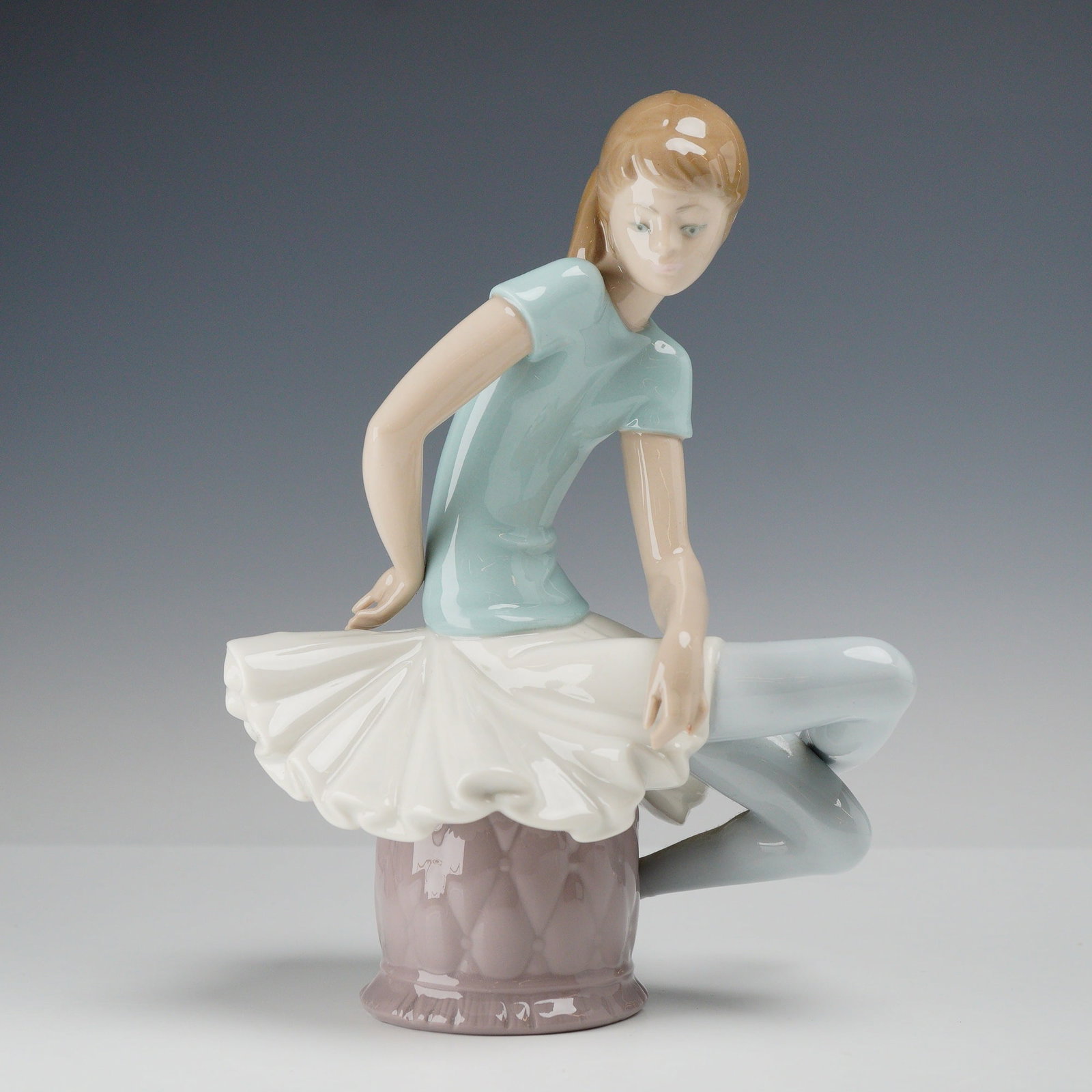Lladro Porcelain Figurine Julia Ballerina Model 1361: Lladro glazed porcelain figurine titled Julia Ballerina, model number 1361. The figurine depicts a young ballerina in a seated, graceful pose on a mauve-colored circular ottoman with a tufted, quilted