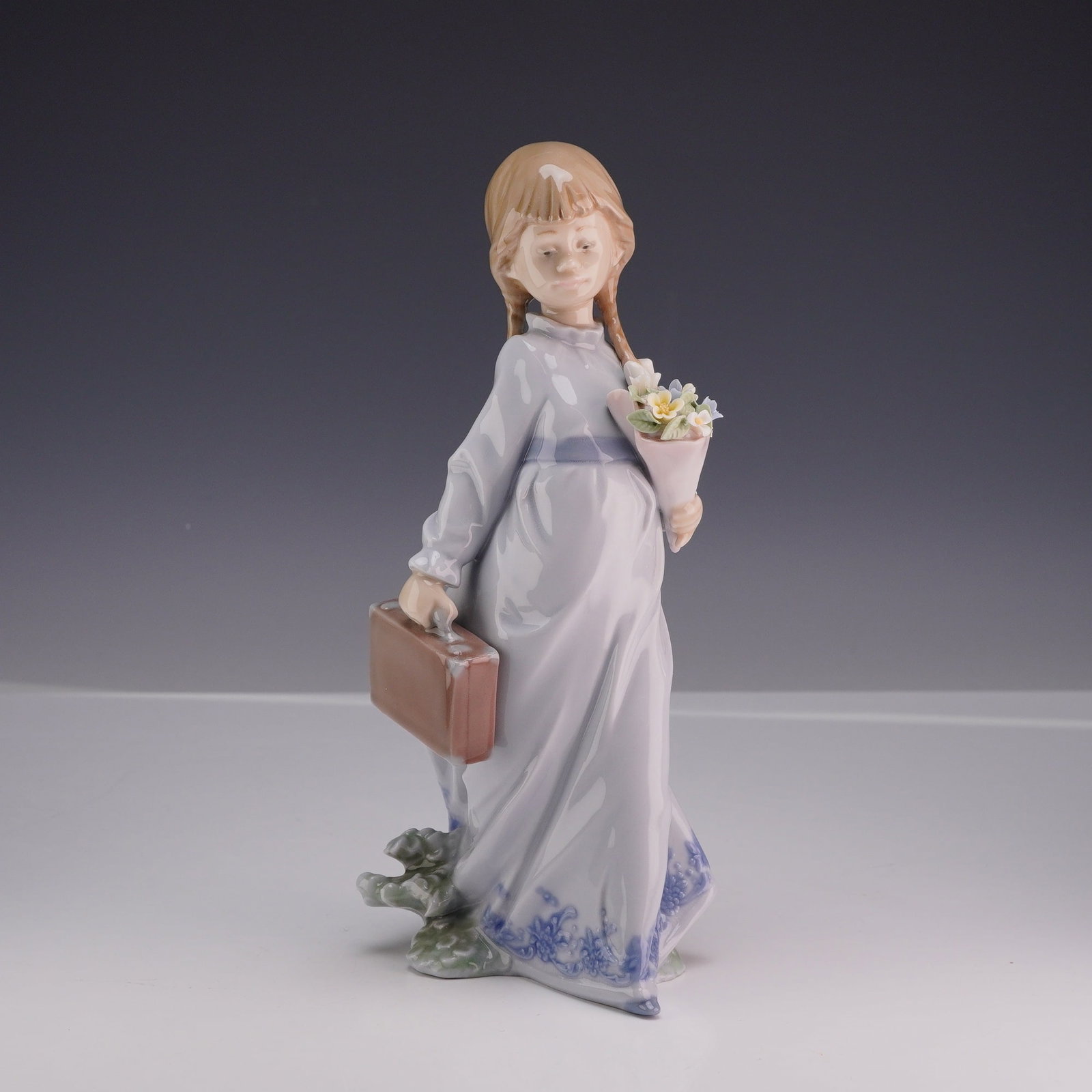 Lladro Porcelain Figurine School Days 01007604 with Original Box: A polychrome glazed porcelain figurine by Lladro titled School Days, model number 7604. The figurine depicts a young girl with blonde hair tied in pigtail braids, wearing a long periwinkle blue dress 