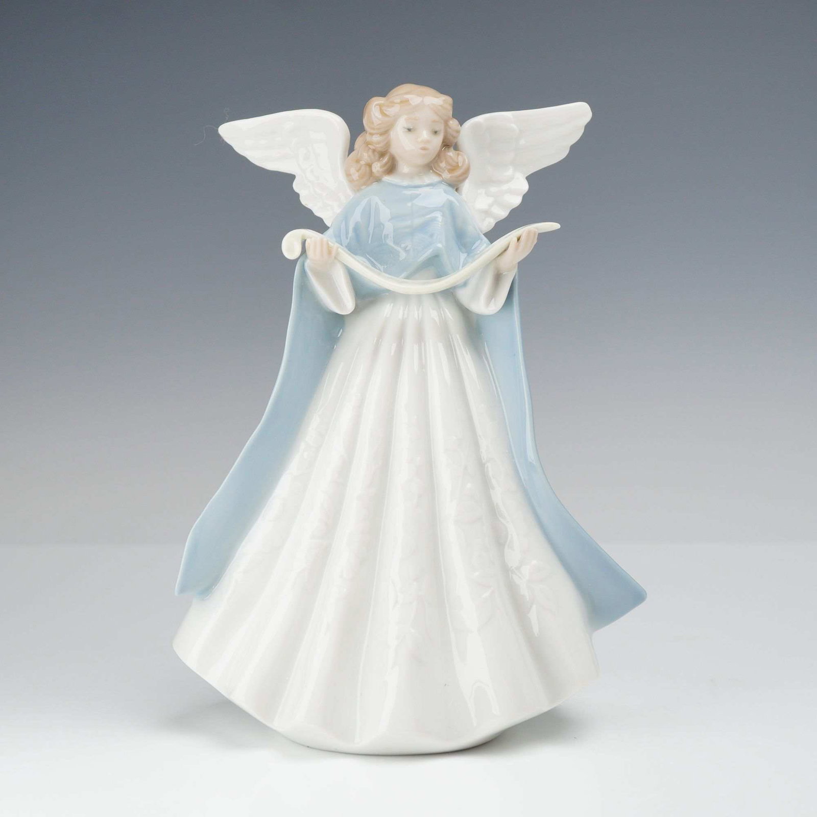 Lladro Porcelain Figurine Tree Topper 1990 (Blue) 01005719: This Lladro glazed porcelain figurine depicts a standing angel with blonde hair and outspread wings. The figure is dressed in a white pleated gown and a long light blue cloak, holding a white scroll i