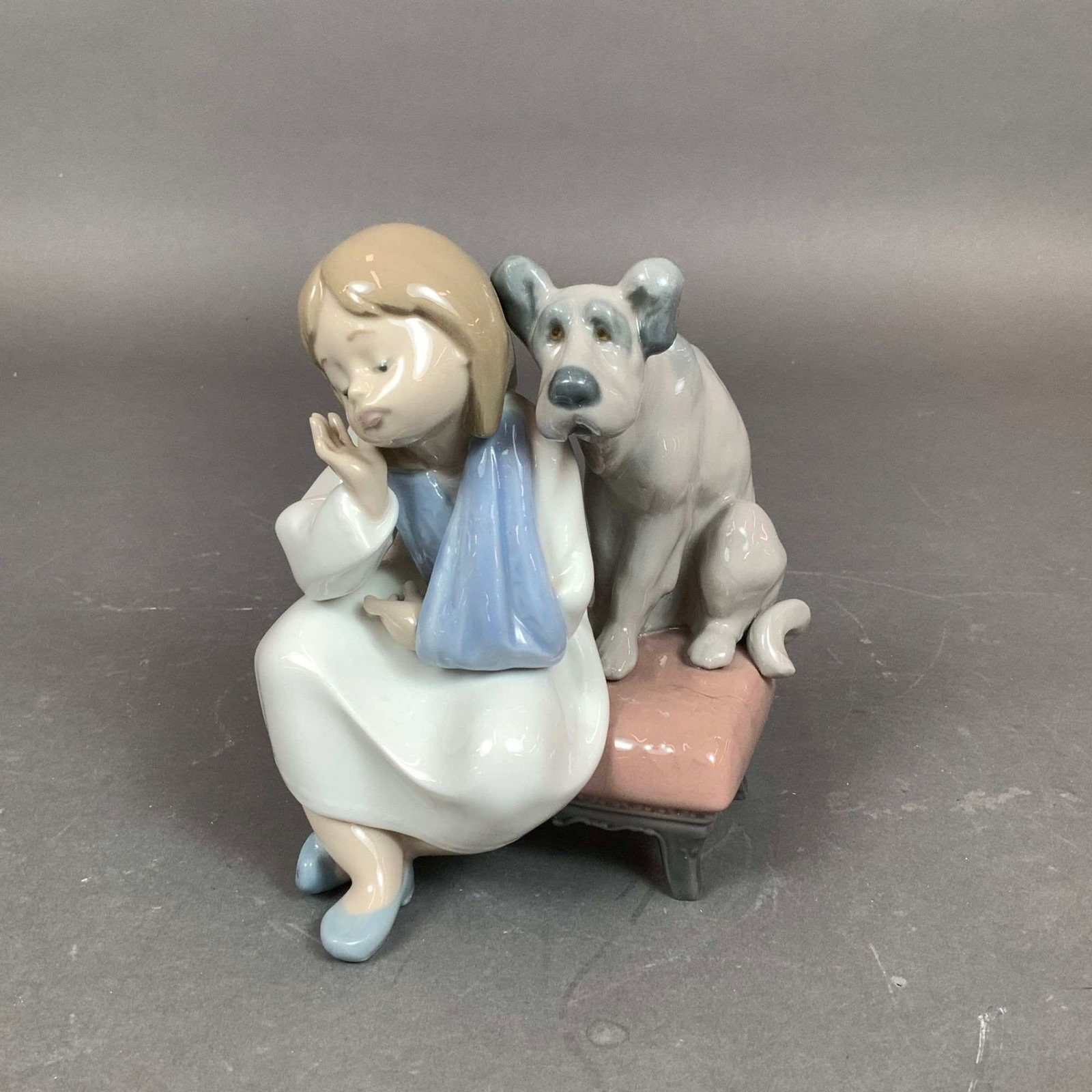 Lladro Porcelain Figurine We Can't Play 01005706: This glazed porcelain figurine, titled A Friend for Life (Spanish title: Un Amigo Inseparable), depicts a young girl dressed in a white nightgown with her left arm held in a light blue sling. She is s